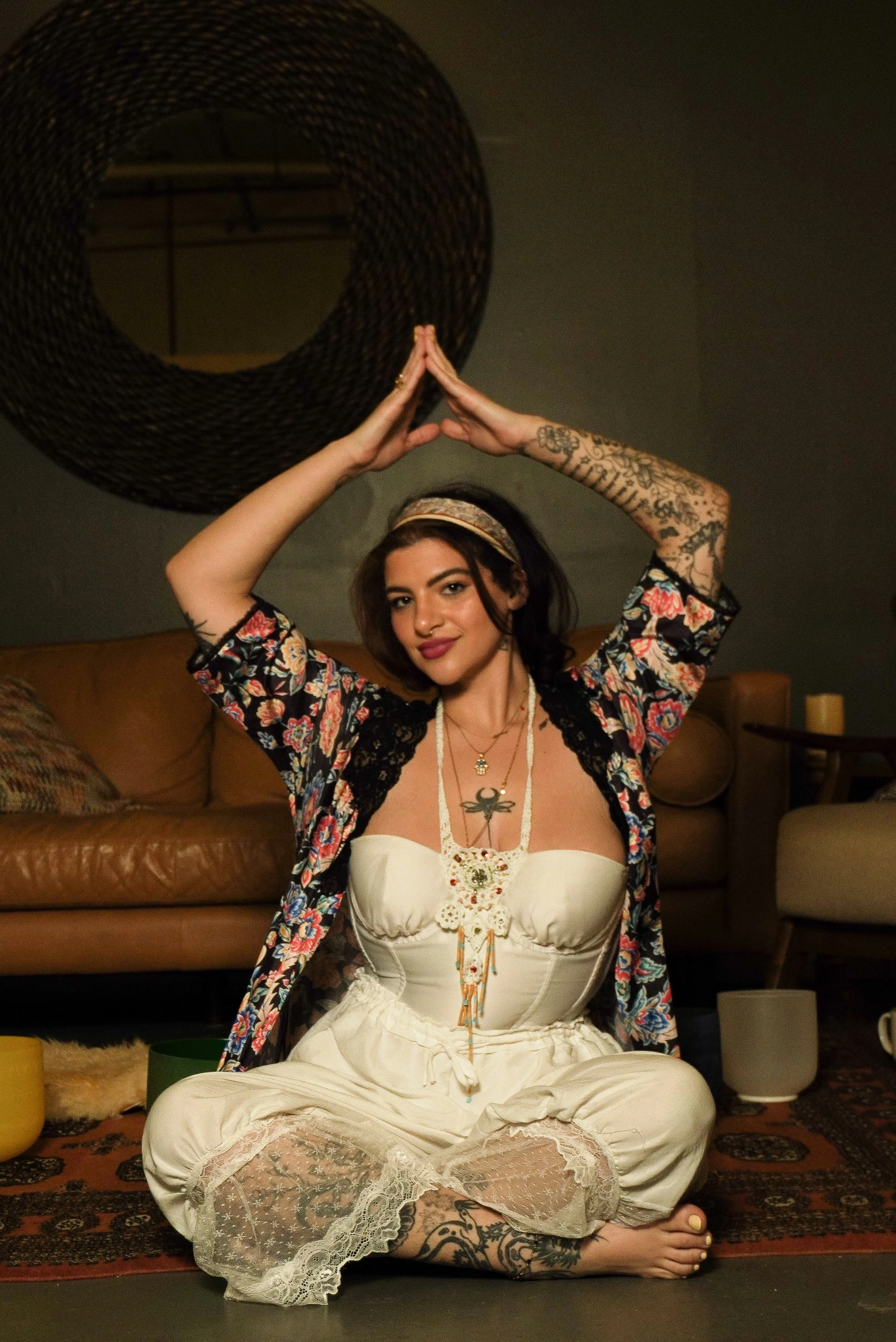 A woman with tattoos and dark hair seated cross-legged on a patterned rug, wearing a bohemian outfit, with her hands raised above her head in a prayer pose in a dimly lit room.