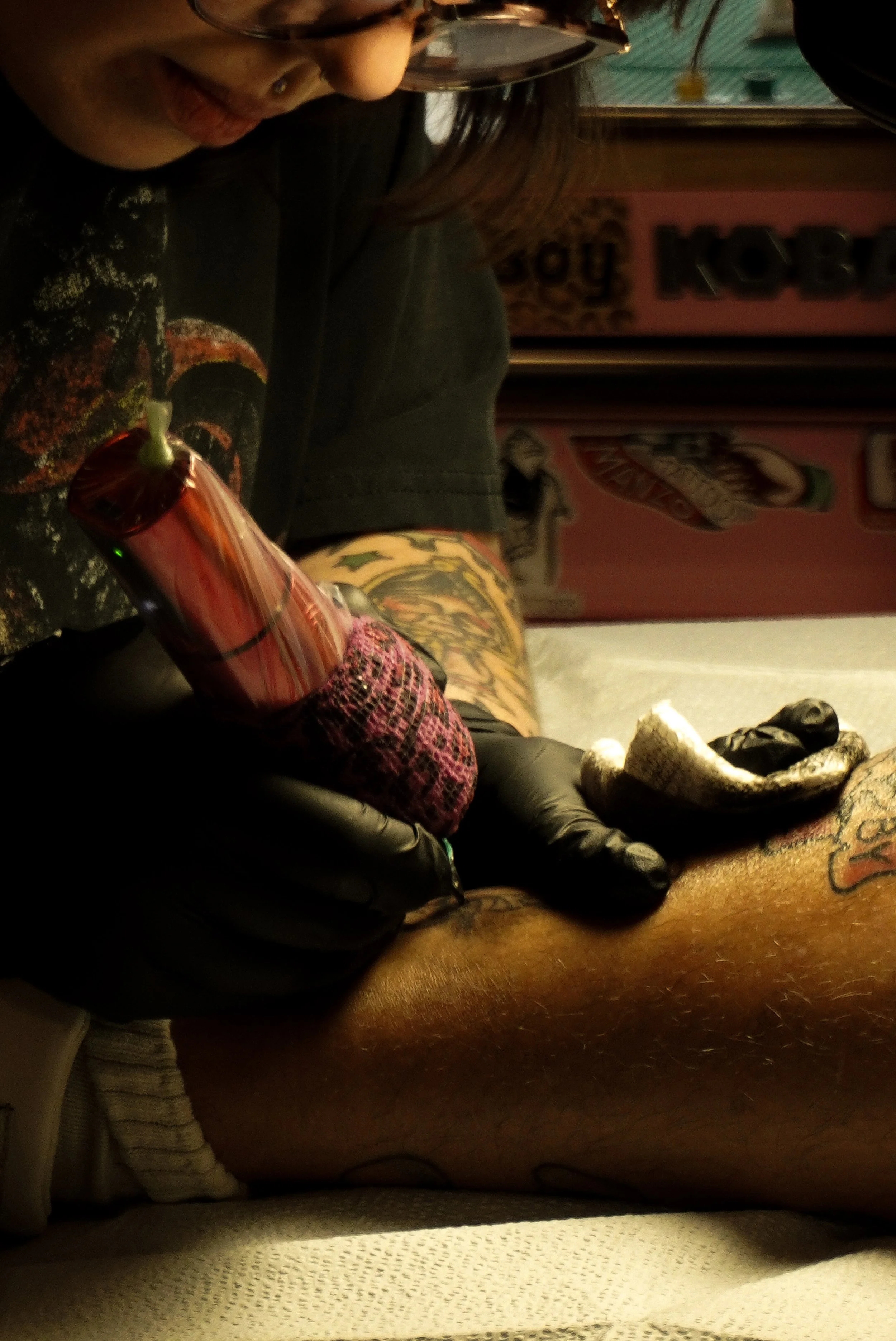 A person getting a tattoo on their leg in a tattoo studio. The tattoo artist is working with ink and a tattoo machine, wearing gloves. The person’s leg is tattooed with colorful designs, and the person is wearing glasses and a black shirt with a graphic design.