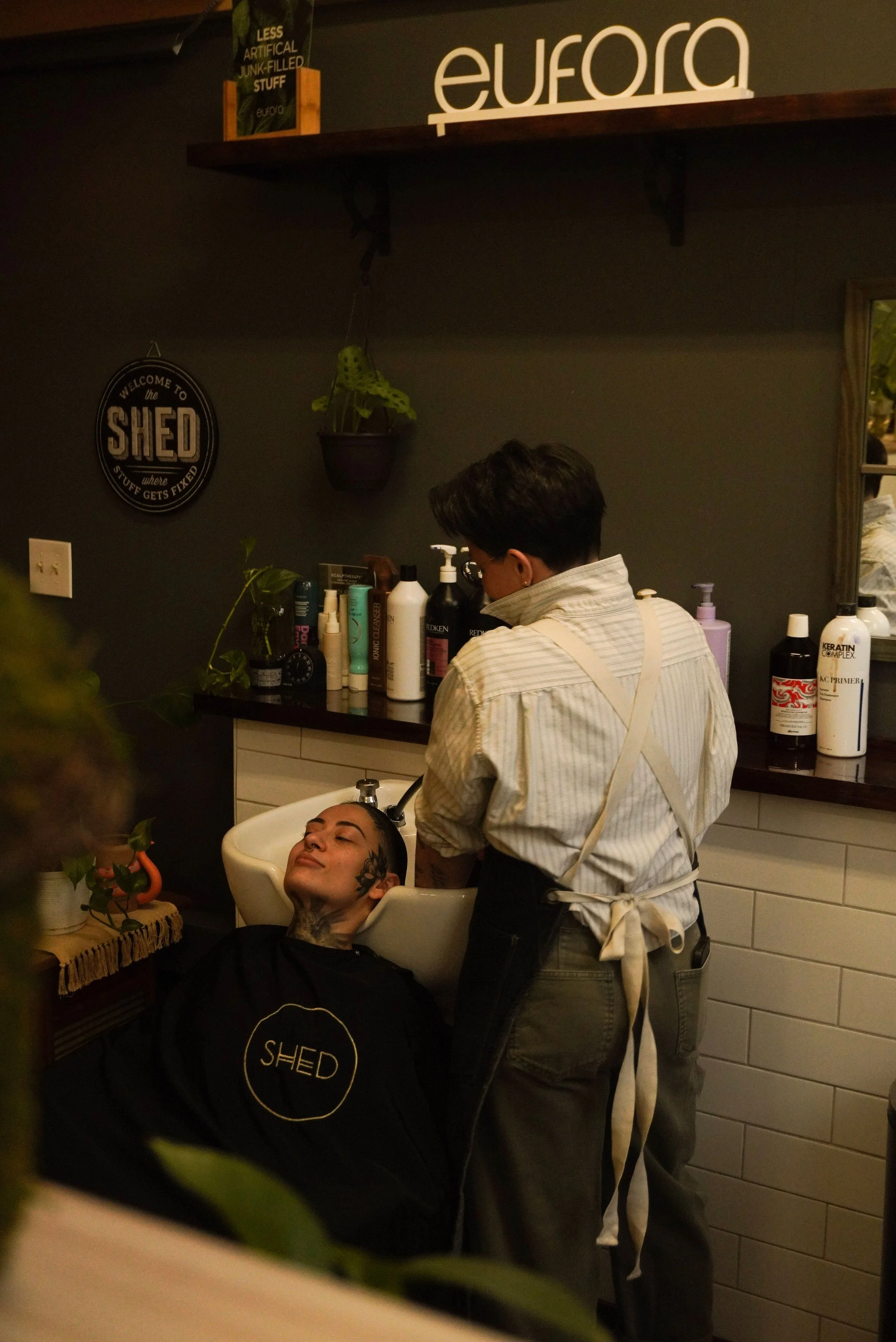 A person with tattoos lying back in a salon chair, getting their hair washed, with a hairstylist standing nearby. The salon has dark-colored walls, a shelf with hair products, and a decorative sign on the wall.