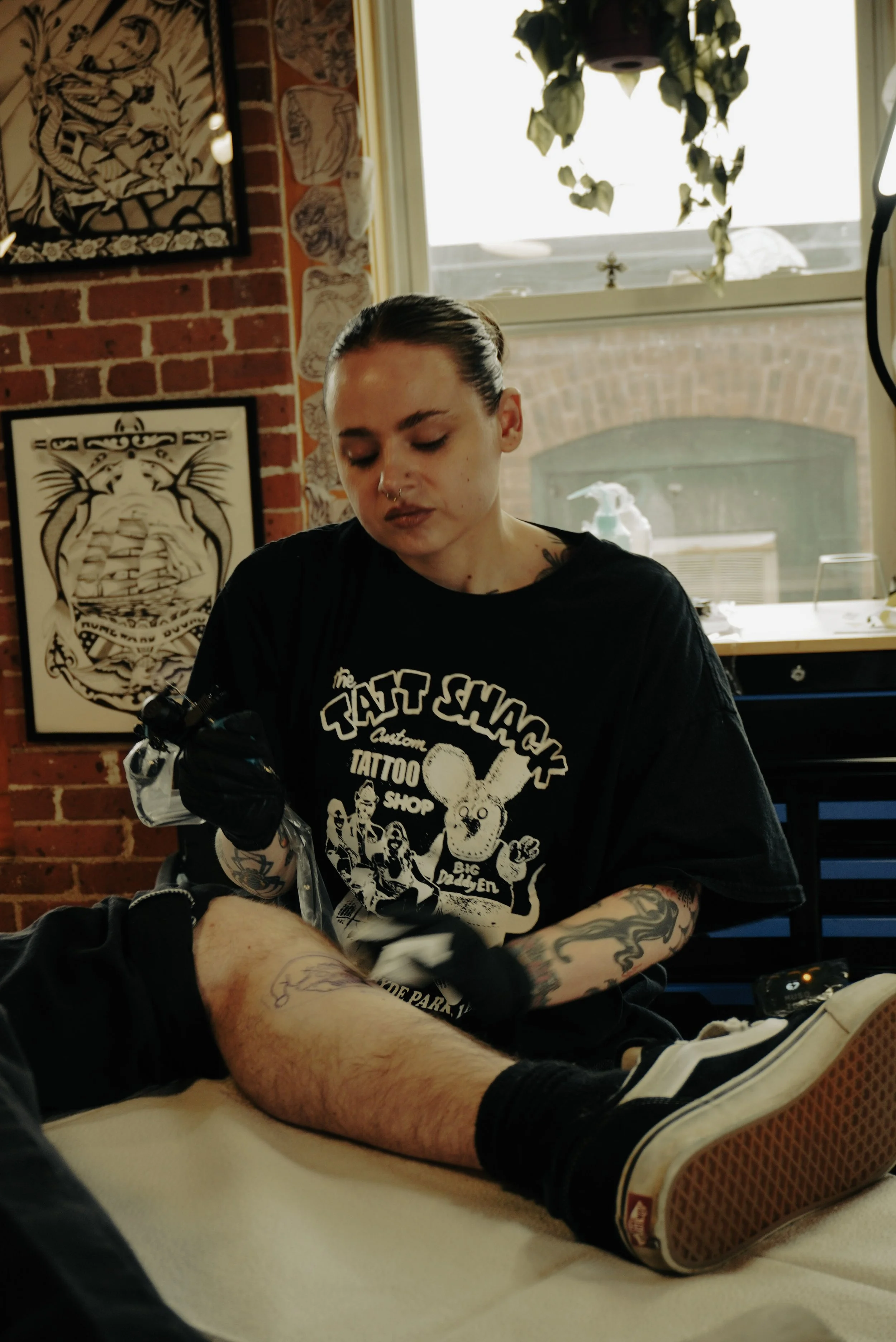 A woman with tattoos on her arms sitting in a tattoo shop, tattooing a person's leg.