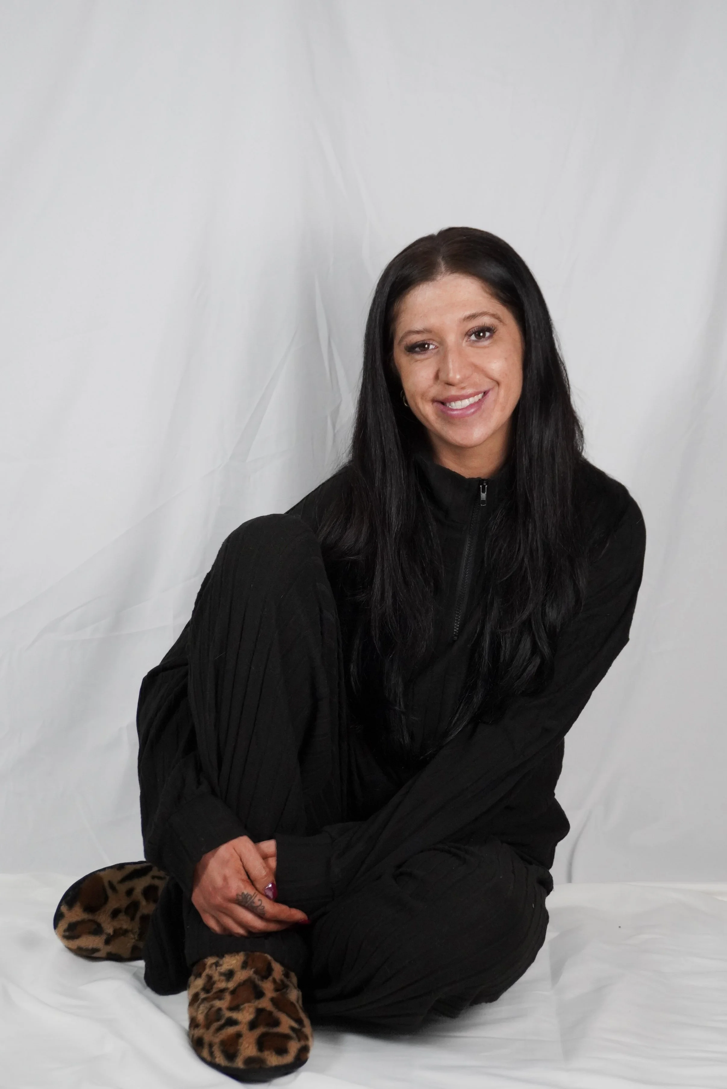A woman with long black hair smiling, sitting on a white surface against a white backdrop, wearing black clothing and leopard print slippers.