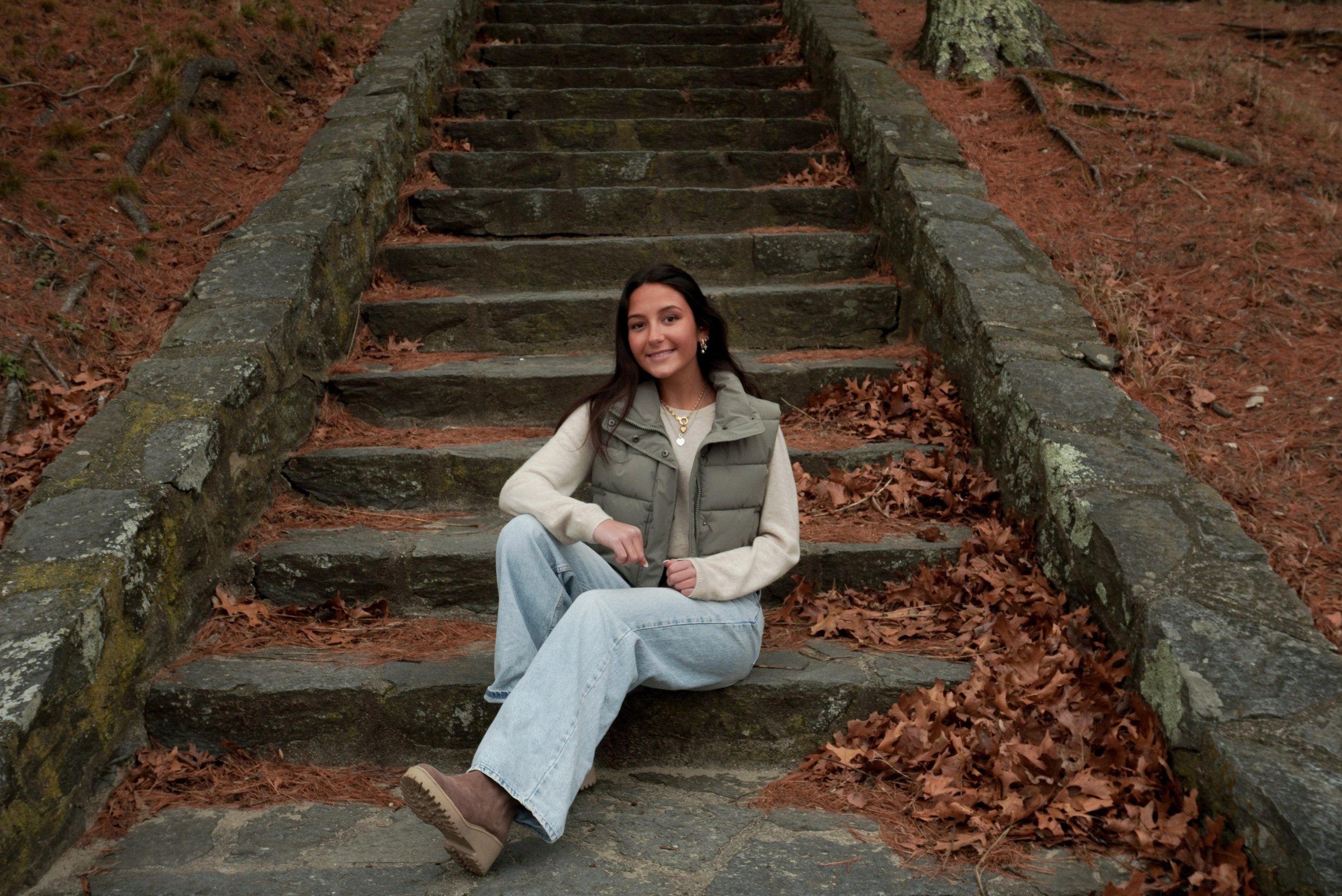 A young woman with dark hair, wearing a beige sweater, gray vest, and light blue jeans, sits on stone steps in an outdoor wooded area with fallen leaves around her.