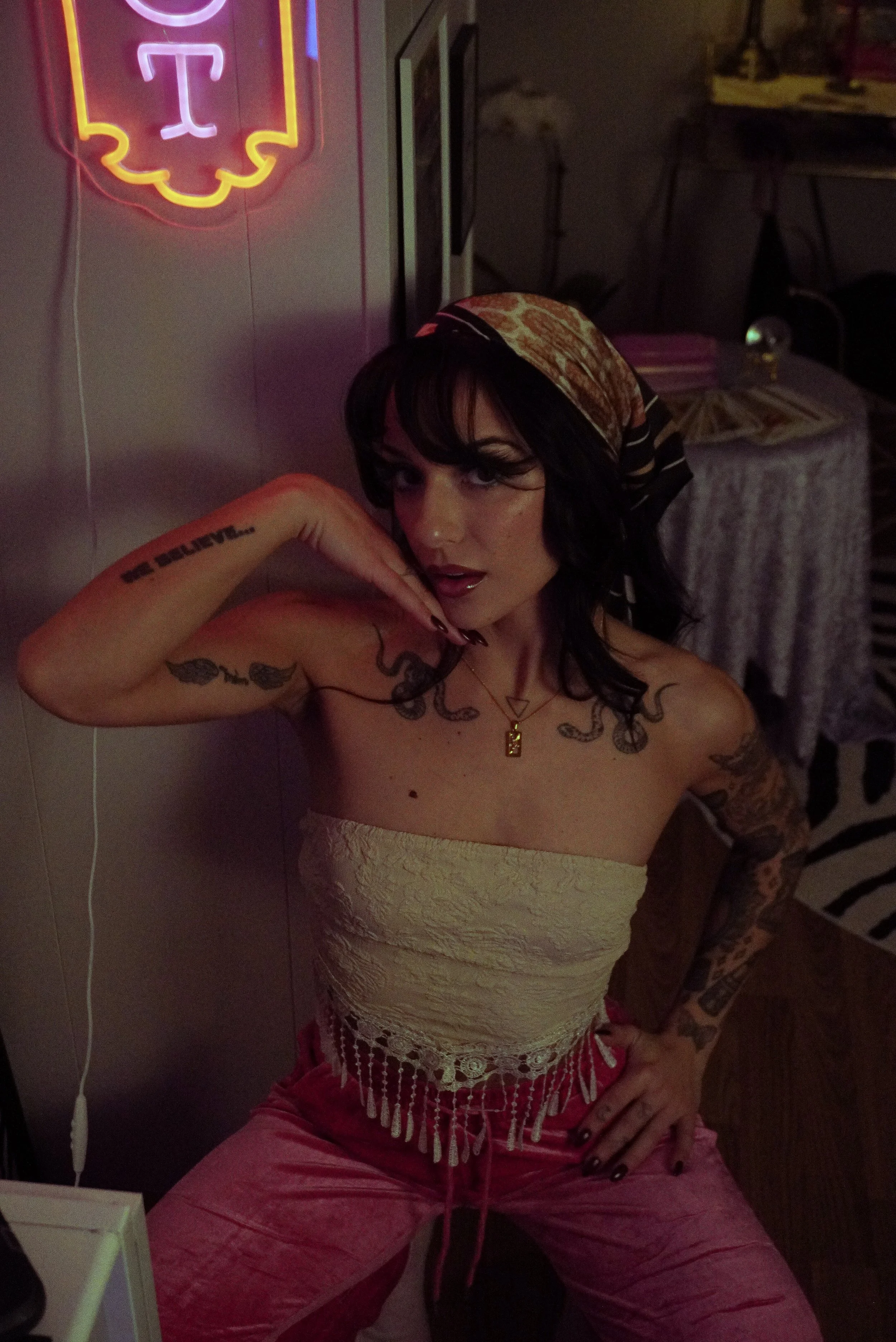 Woman with tattoos wearing a headscarf, beige strapless top, and pink pants, posing with one hand on her hip and the other near her face in a dimly lit room with neon signs.