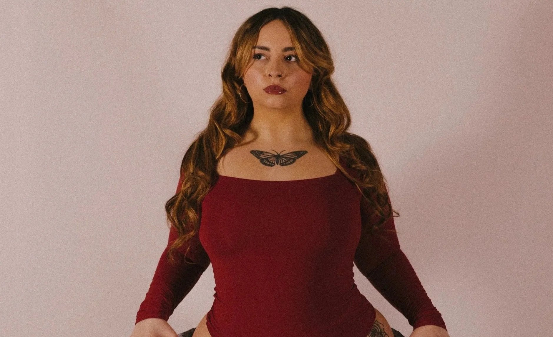 A woman with long, curly brown hair and a butterfly tattoo on her chest, wearing a fitted red long-sleeve top, looking to the side against a plain background.