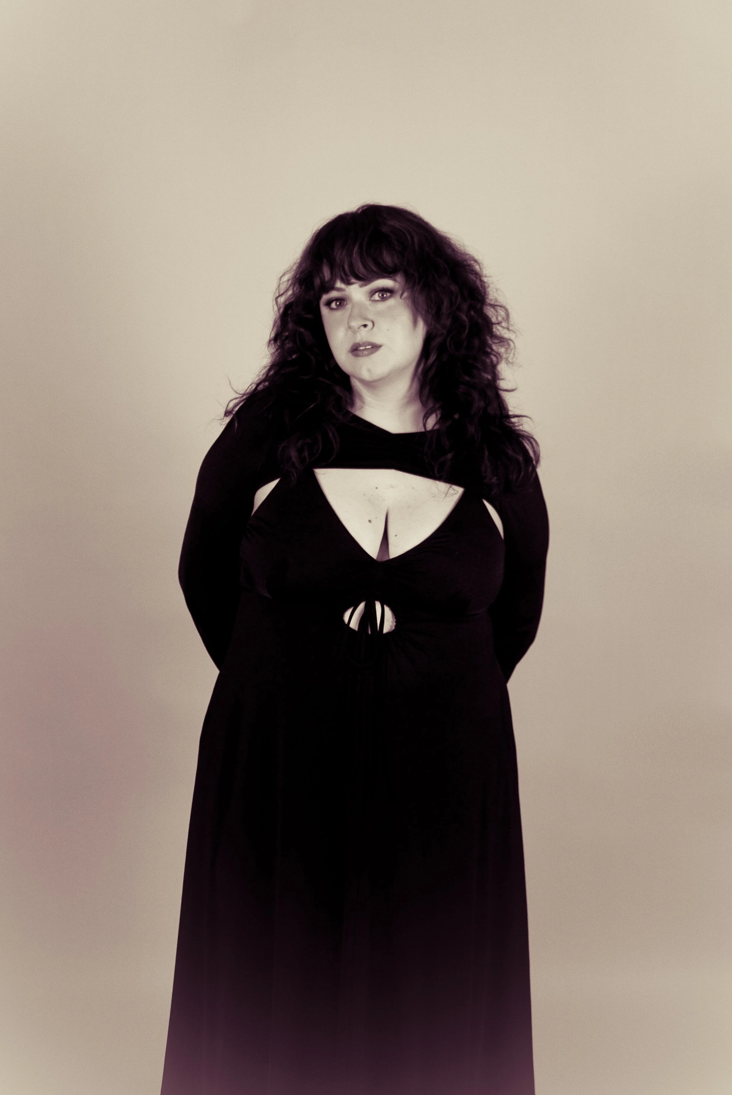 Woman with curly dark hair wearing a long black dress with cutouts, standing against a beige background.