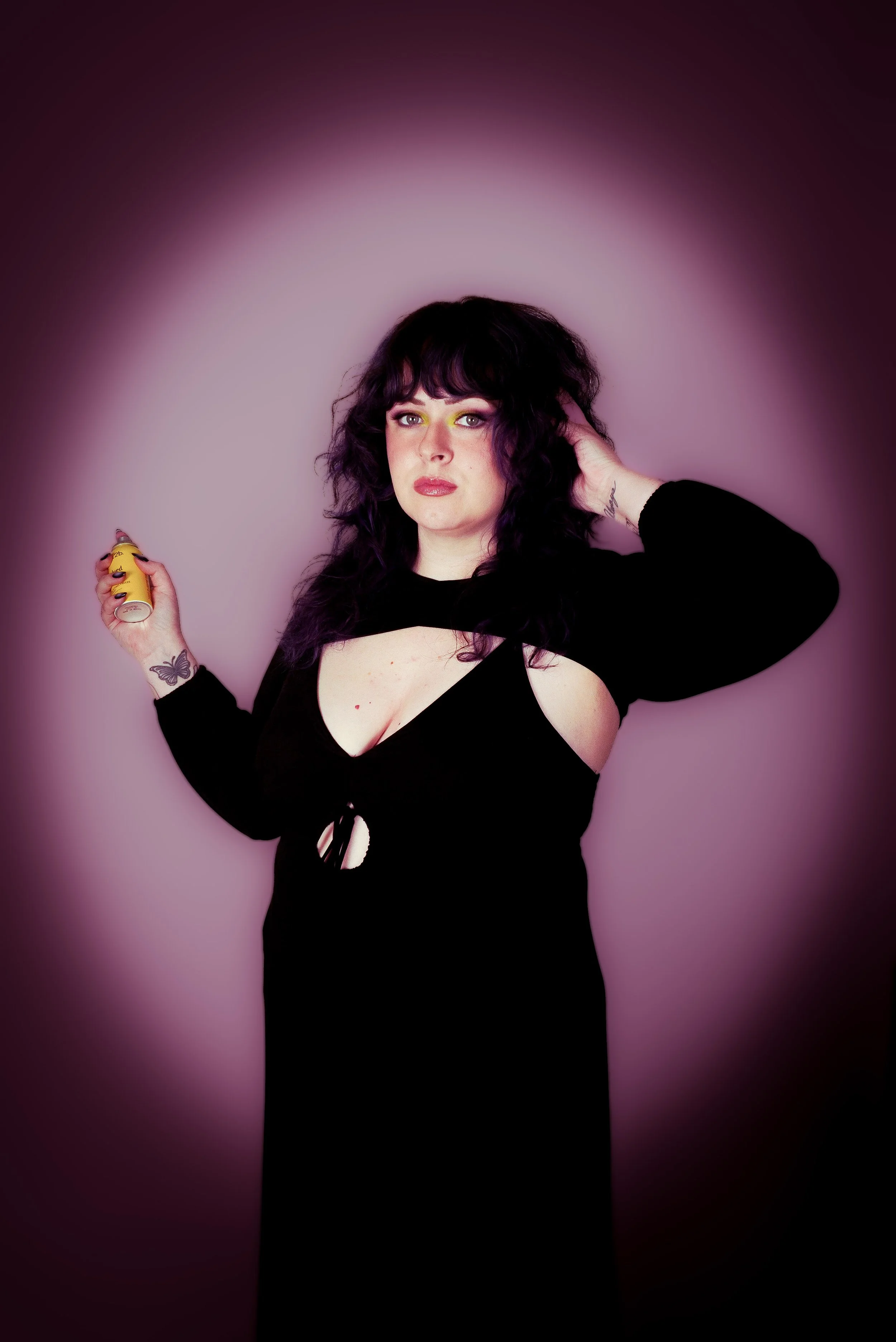 A woman with curly black hair and tattoos wearing a black dress with cutouts, holding a yellow spray paint can, posing against a pink gradient background.