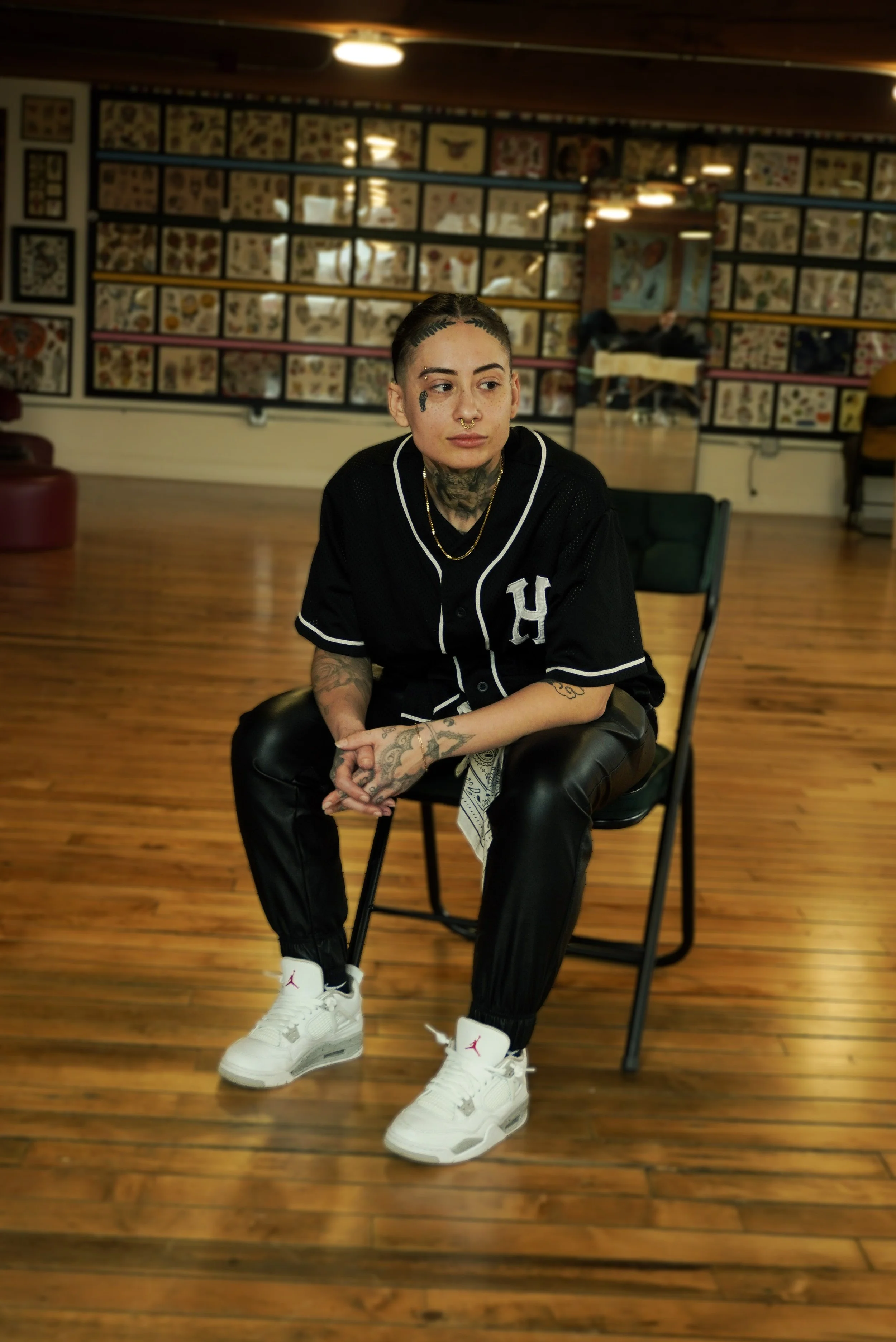 A young woman with tattoos, dark hair, and multiple piercings, wearing a black baseball jersey, white sneakers, and sitting on a black chair in a room with wooden floors and tattoo flash art on the walls.
