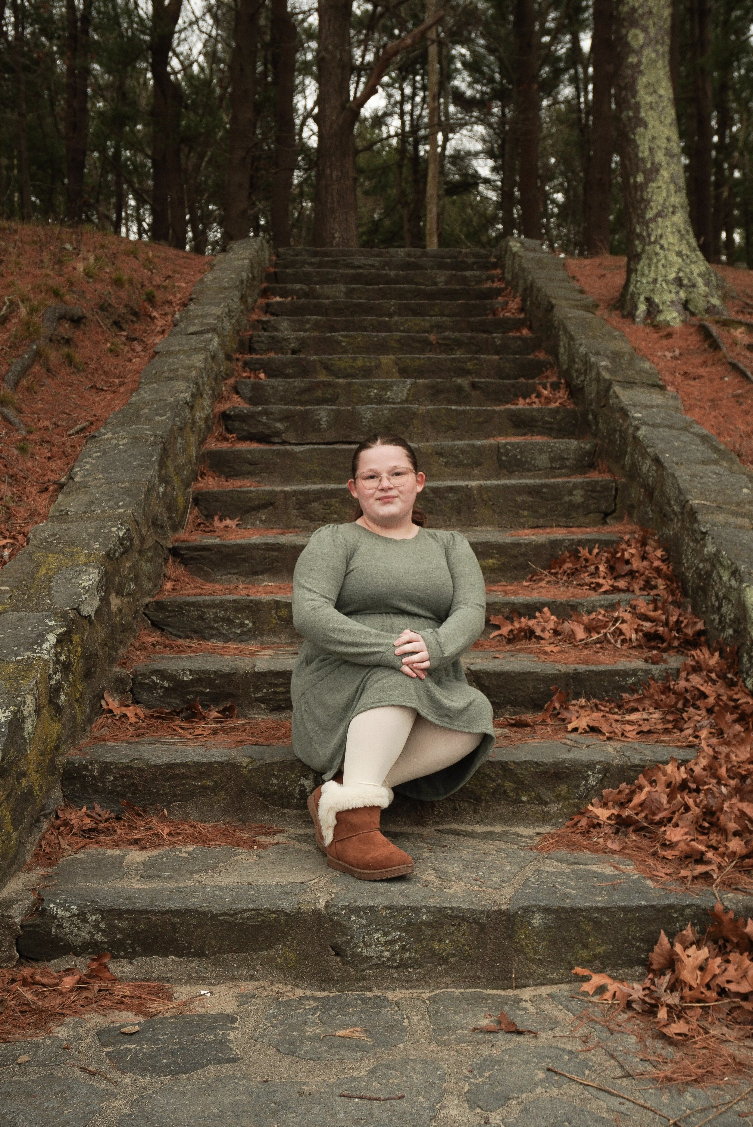 A woman in a green dress, white tights, and brown boots sitting on stone steps outdoors surrounded by trees and fallen leaves.