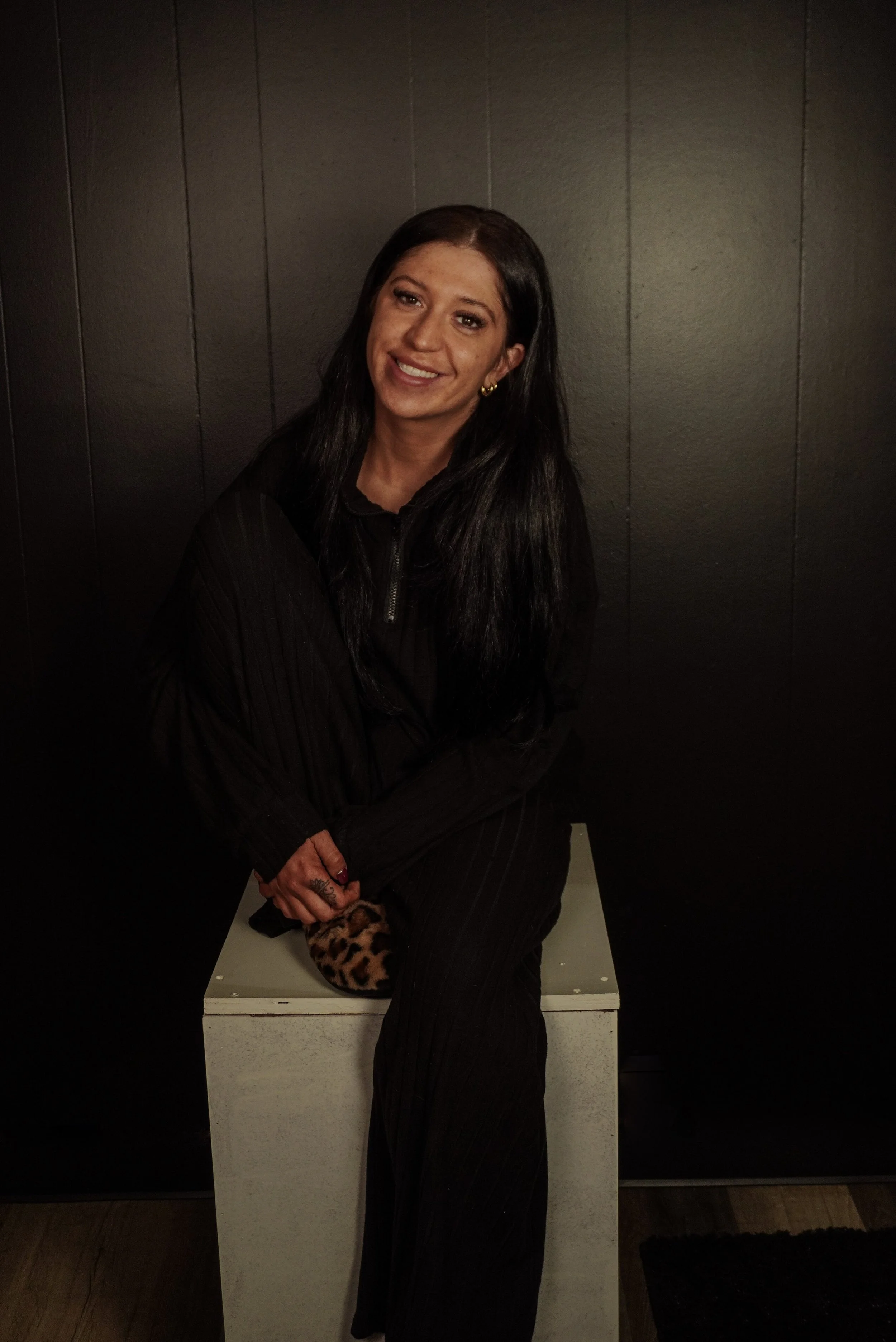 A woman with long dark hair sitting on a white cube, smiling, wearing a black outfit, and holding a leopard-print bag.