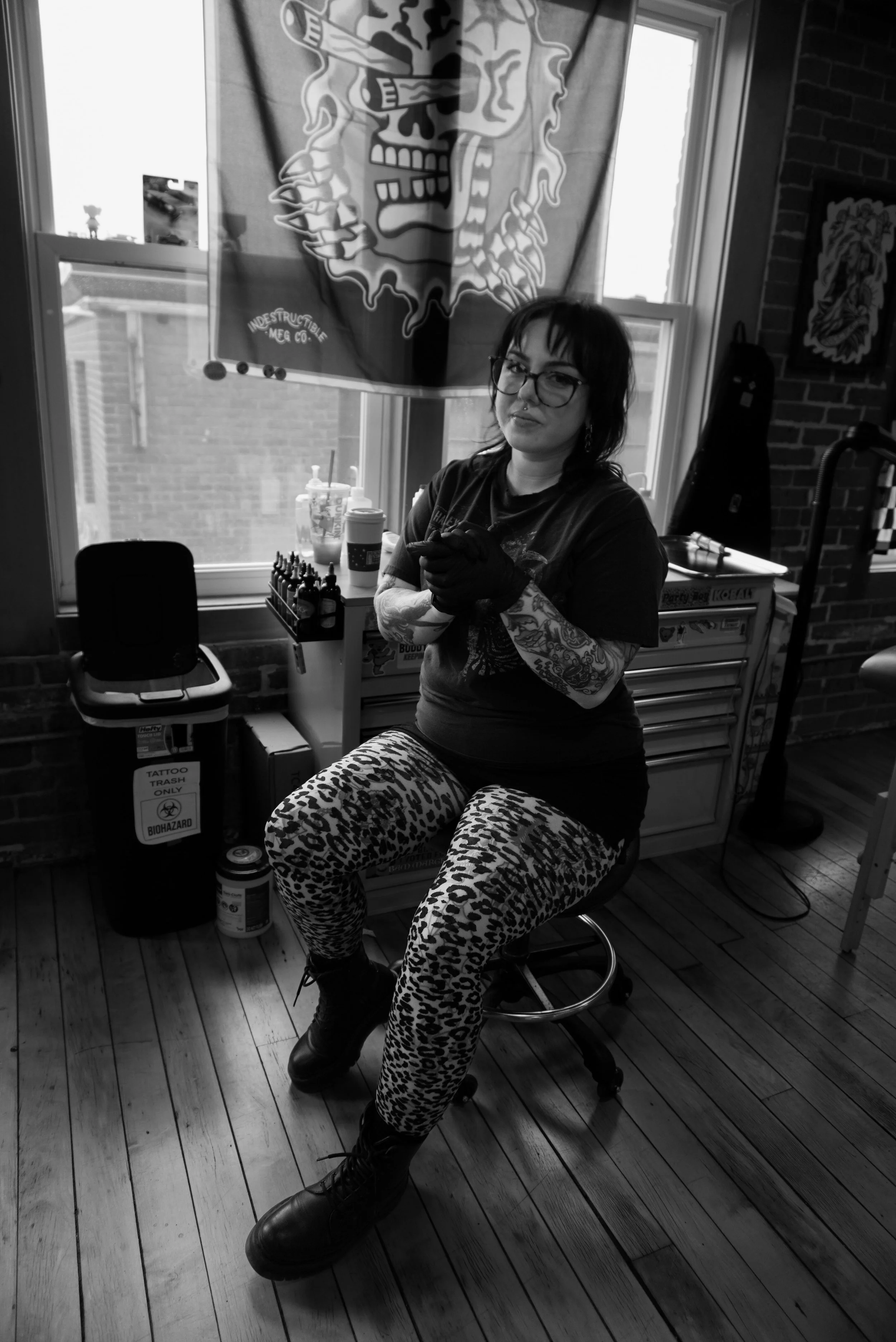 A woman with glasses, tattoos, and leopard print pants sitting on a stool in a tattoo studio, with tattoo supplies and trash bin in the background, black and white photo.