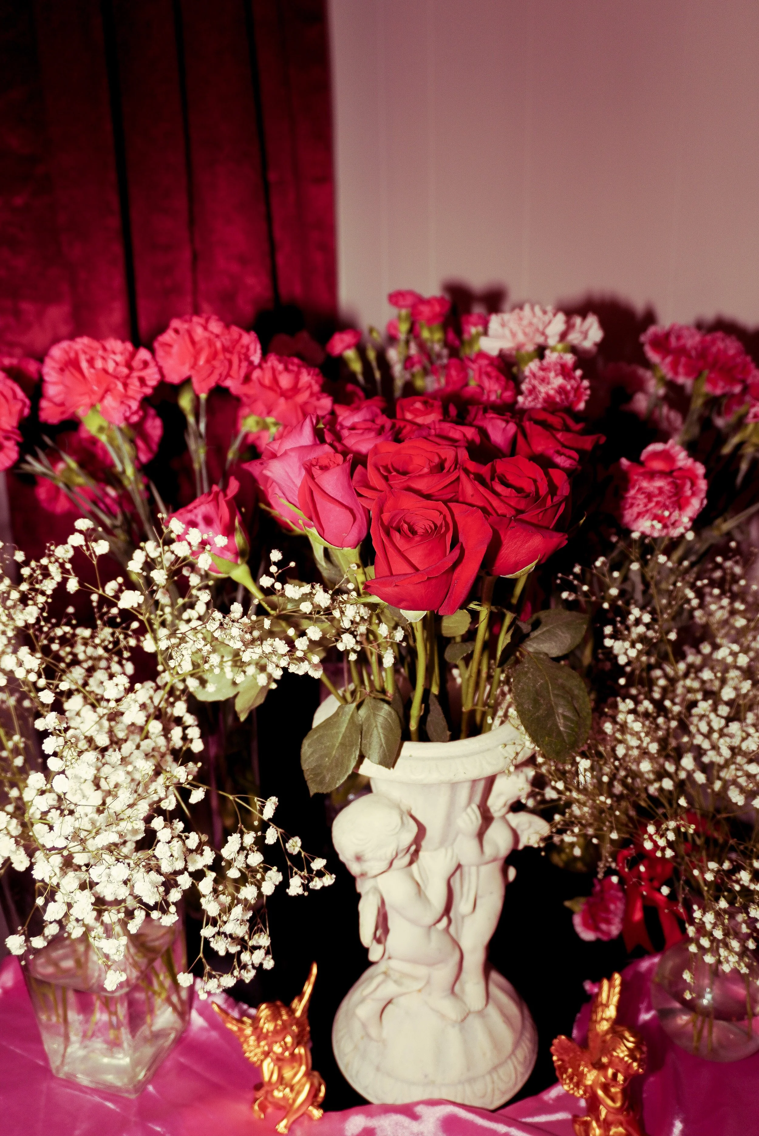 Vase with pink roses, pink carnations, white baby's breath, on pink fabric, with gold animal figurines.