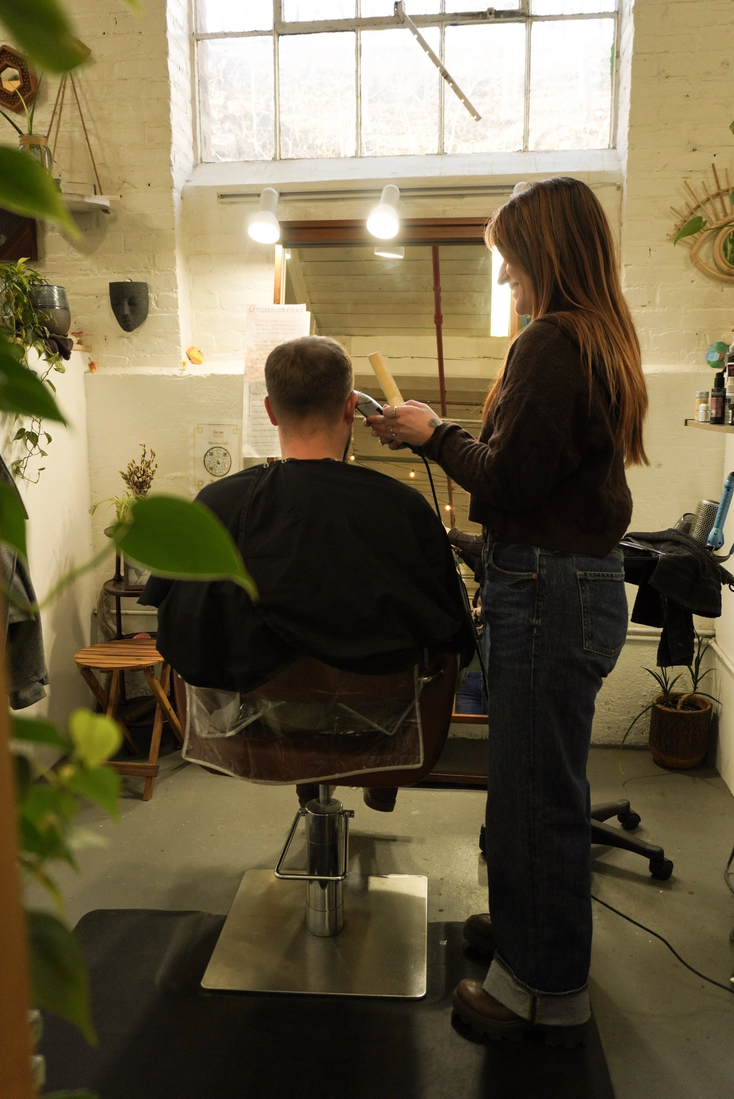 A woman is giving a haircut to a man in a salon with natural and artificial lighting, surrounded by plants and decor.