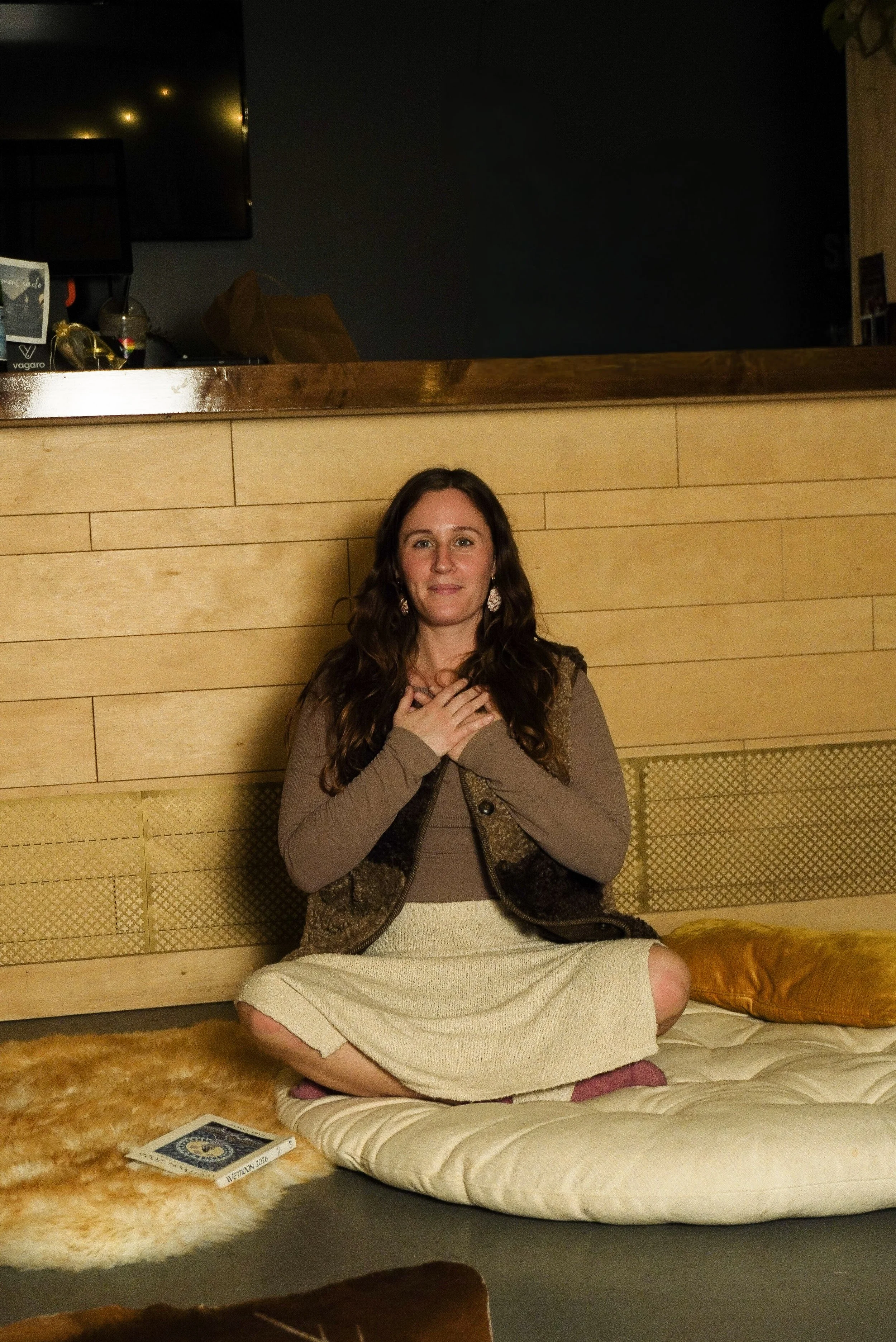 A woman with long brown hair, wearing earrings, a brown top, a multicolored vest, and a beige skirt, is sitting cross-legged on a beige cushion on the floor in a cozy indoor setting. She has a hand on her chest and is looking at the camera, with a warm smile.