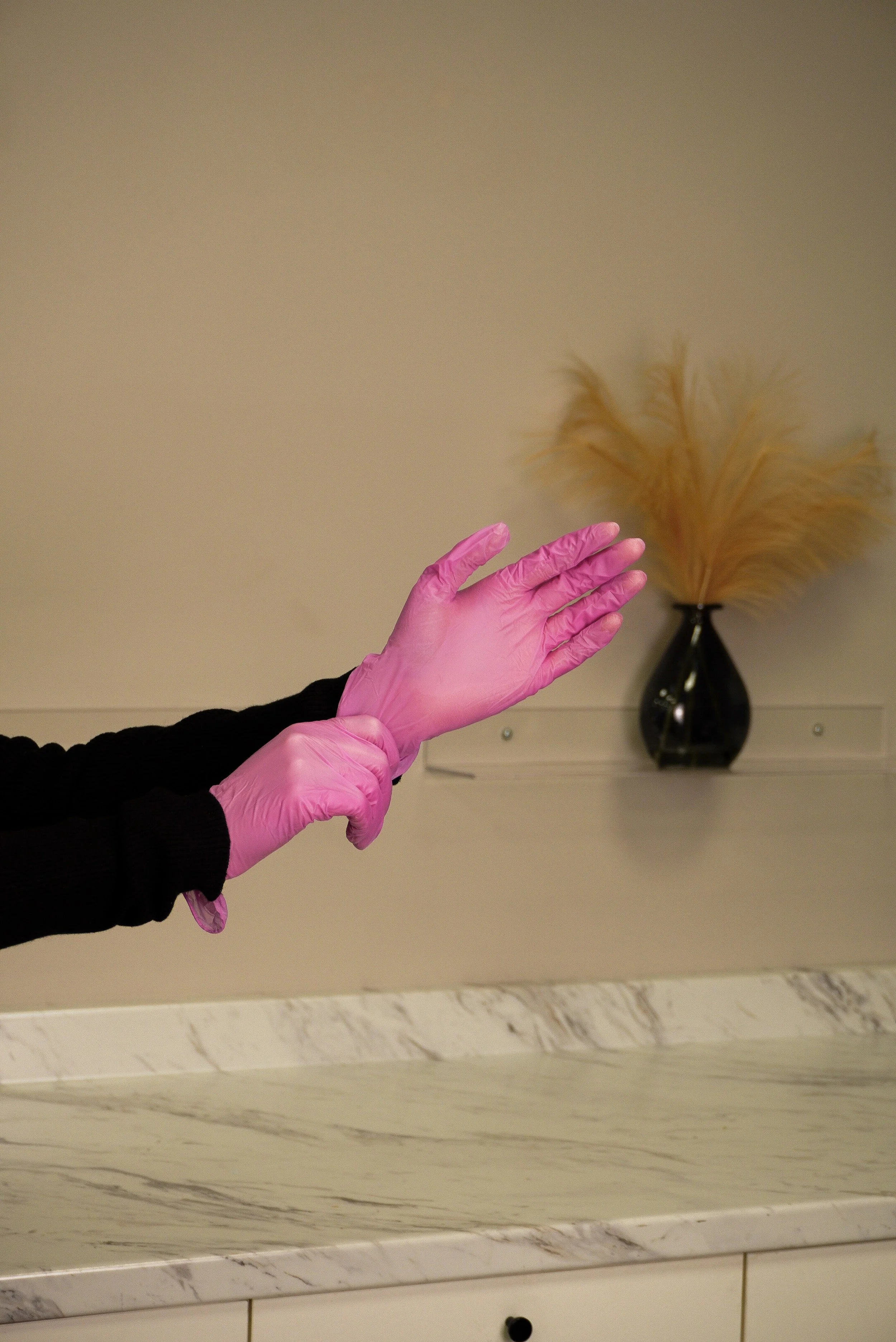 Person wearing pink gloves and a black long-sleeve shirt is stretching or adjusting their gloves in a room with a beige wall and a black vase with beige dried plants on a shelf.