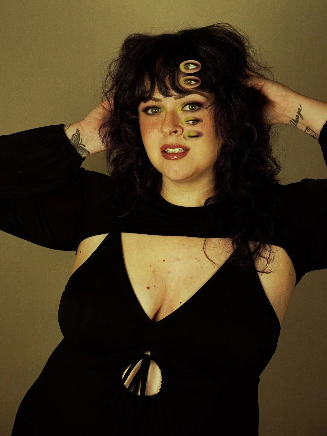 A woman with dark wavy hair and tattoos on her arms poses with her hands behind her head, wearing a black top with a cut-out section and multiple eye-shaped objects covering parts of her face.