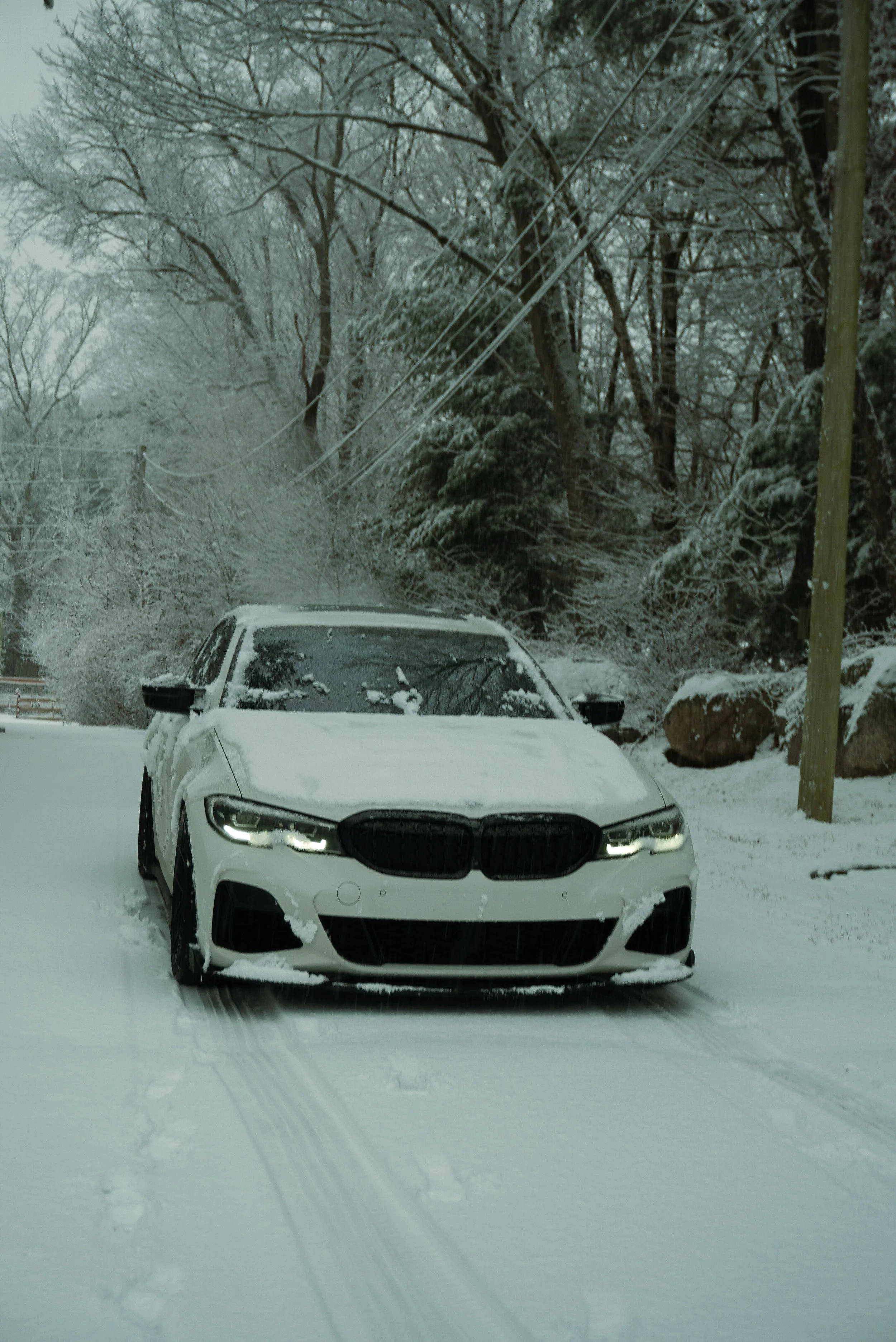 A white BMW car parked on a snow-covered road surrounded by snow-covered trees and rocks in a winter landscape.