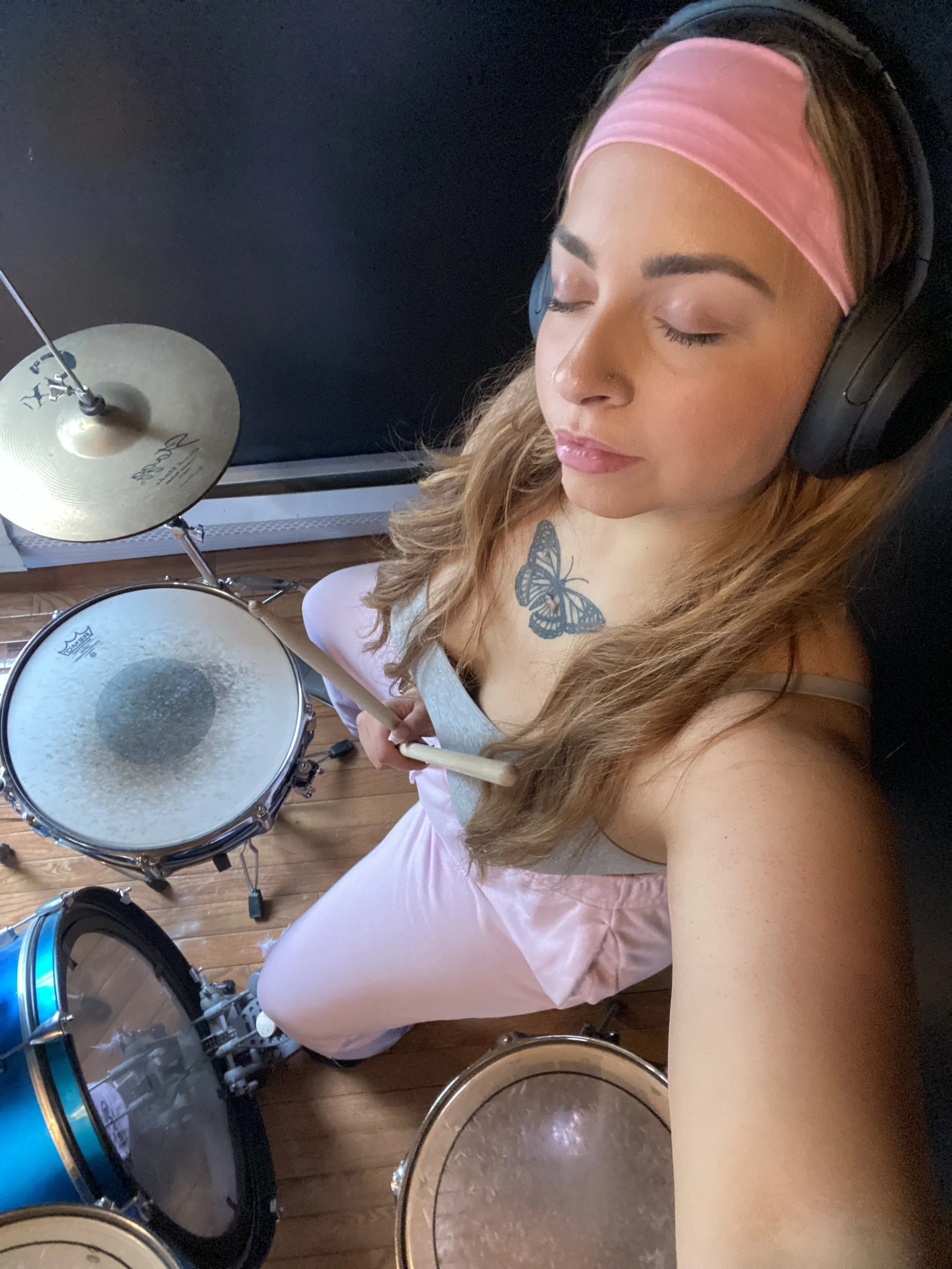 A young woman with a pink headband and large black headphones sitting on the floor with her eyes closed, holding drumsticks over a drum set with a wooden floor below.