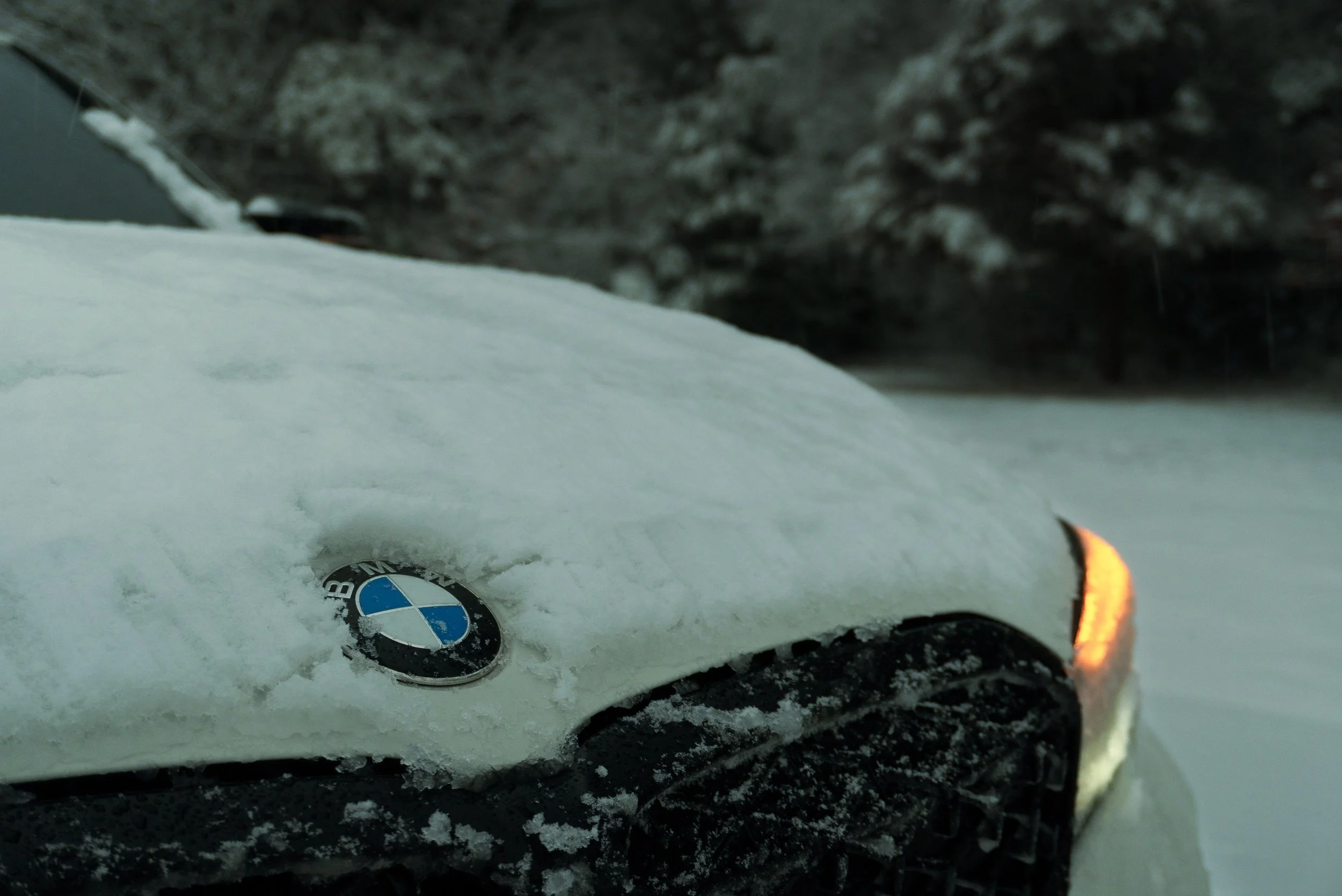 Close-up of a white BMW car covered in snow, with the BMW emblem visible on the hood and the front right orange turn signal on.