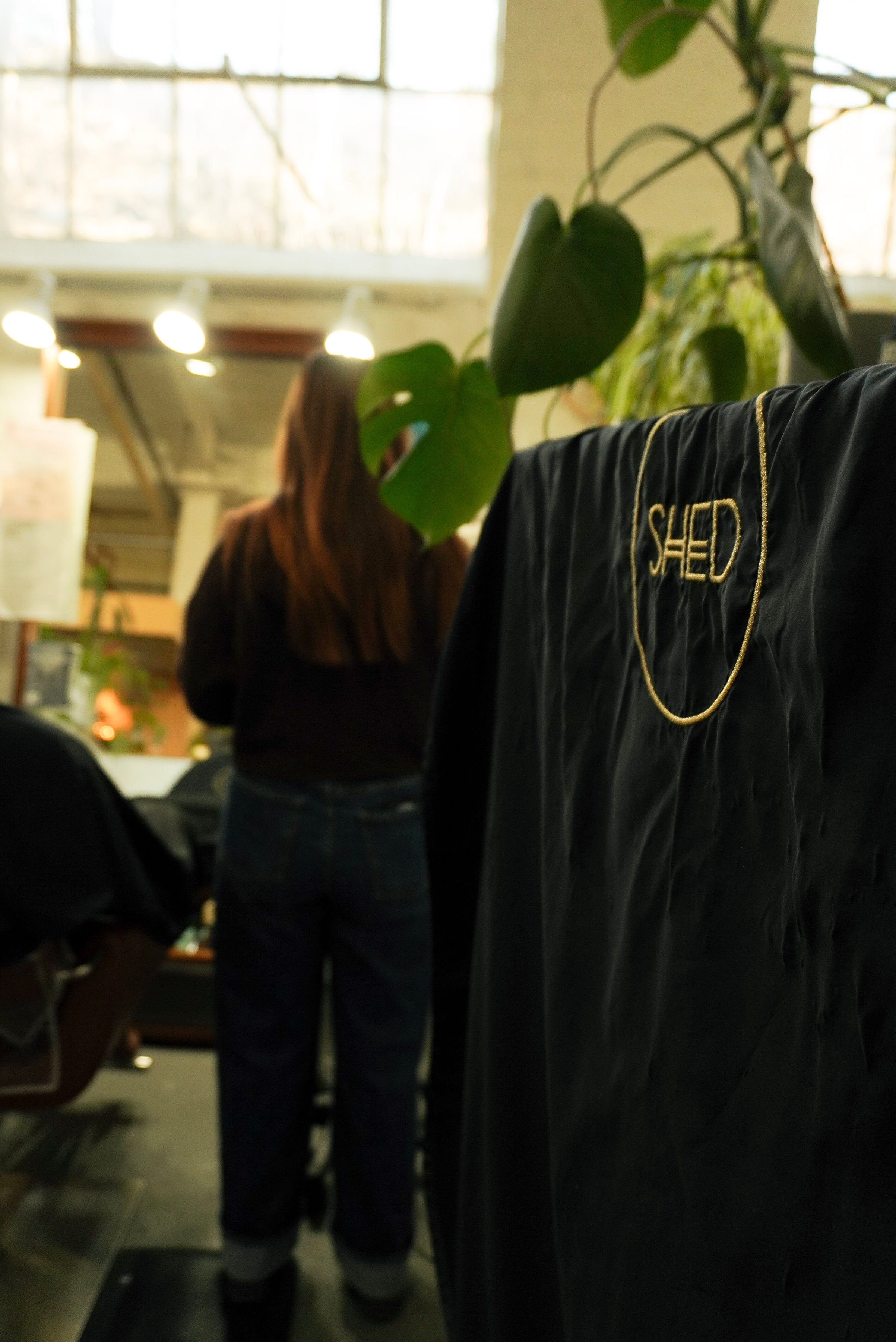 A person with long hair standing in a room with large windows and indoor plants, wearing a black shirt. A black fabric with the word 'SHED' embroidered in gold and a gold chain are in the foreground.
