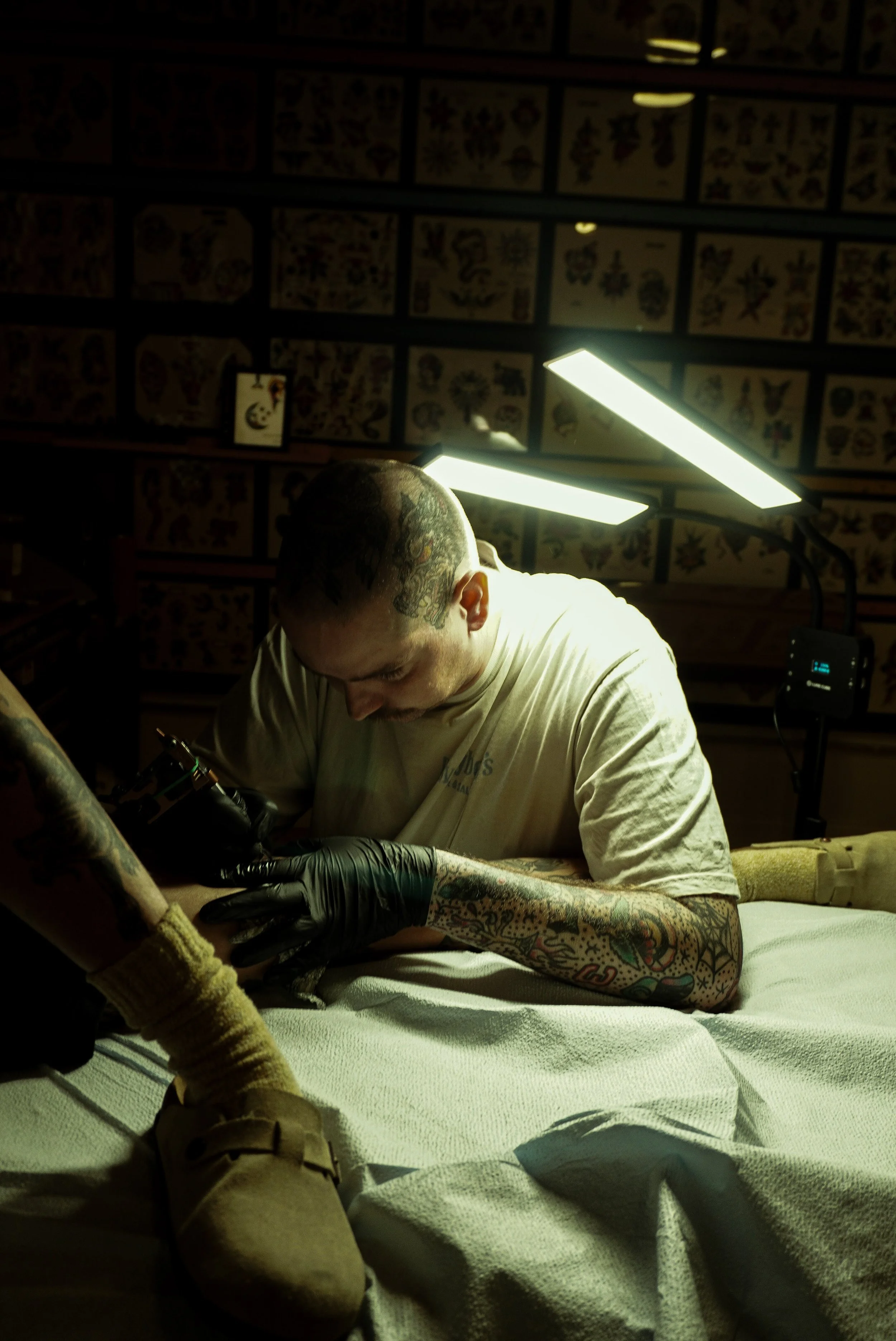 A tattoo artist working on a person's arm in a tattoo studio, with tattoo designs on the wall in the background, illuminated by bright studio lights.