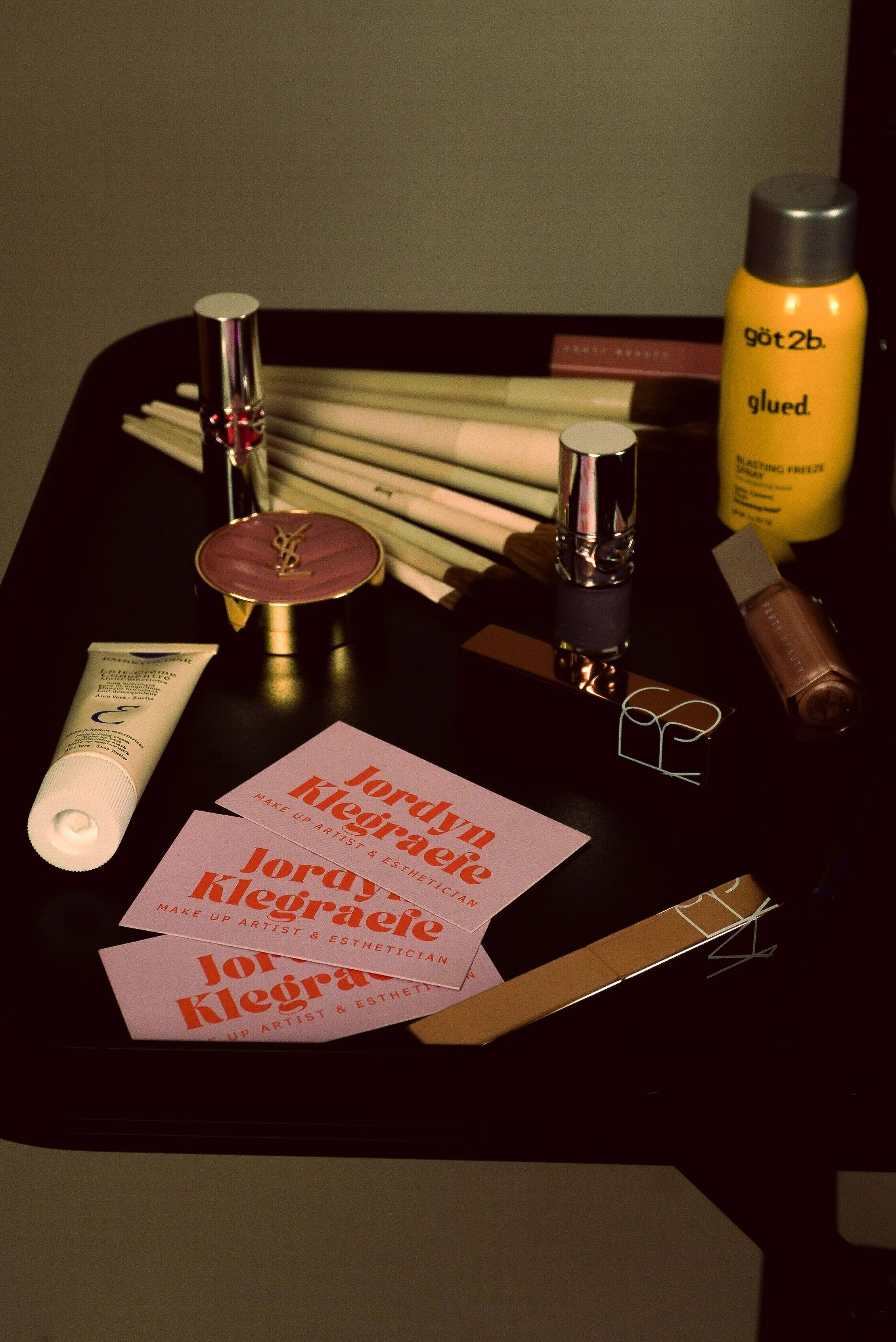 Beauty products, business cards, and hair accessories on a dark table, including Yves Saint Laurent compact, lip gloss, make-up brushes, dry shampoo, and business cards for Jordyn Kearaere, makeup artist and esthetician.
