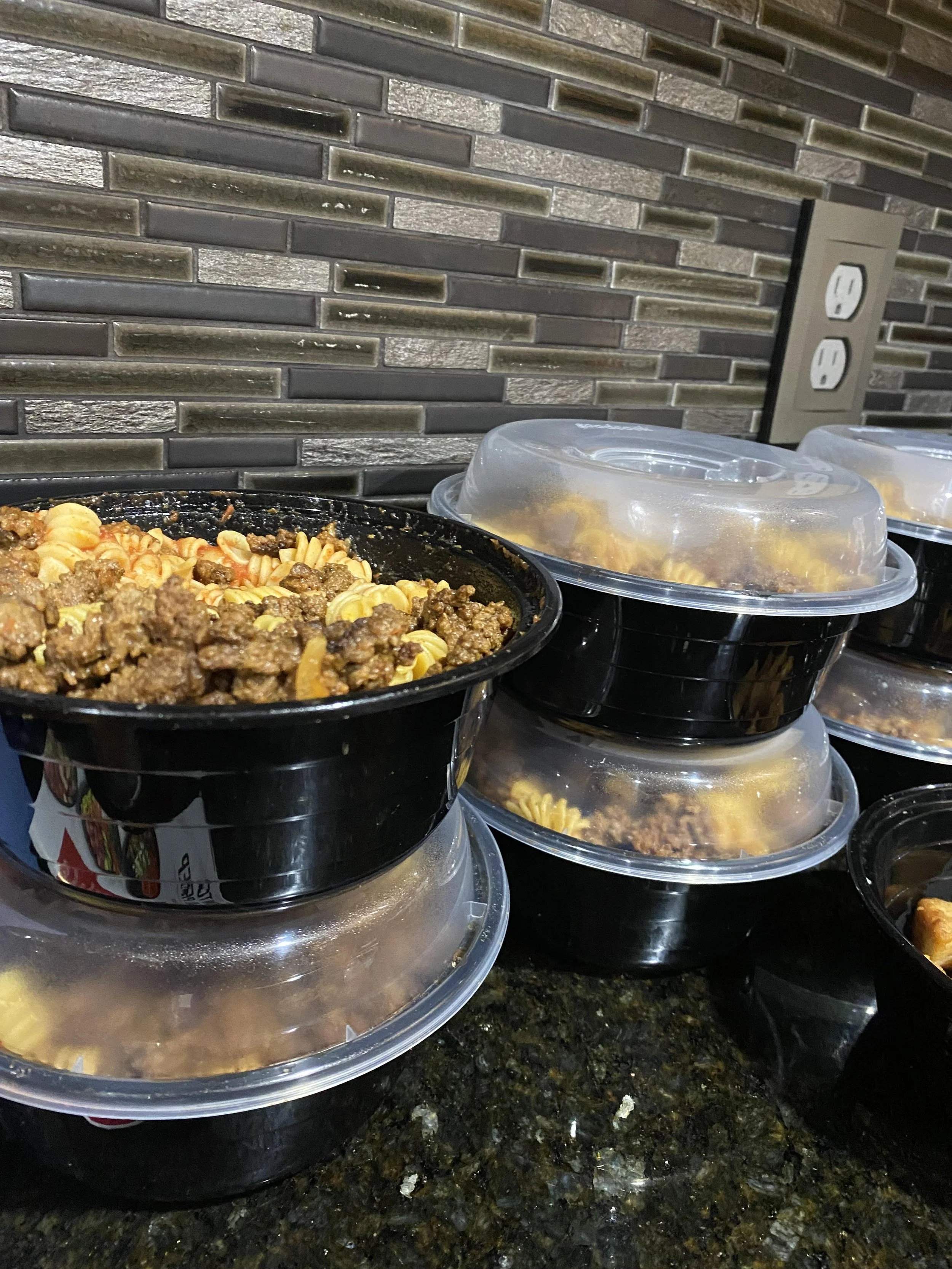 Meal prep containers stacked on a kitchen counter, with cooked pasta and ground beef inside. The containers have clear lids and are placed in front of a textured gray tile backsplash.