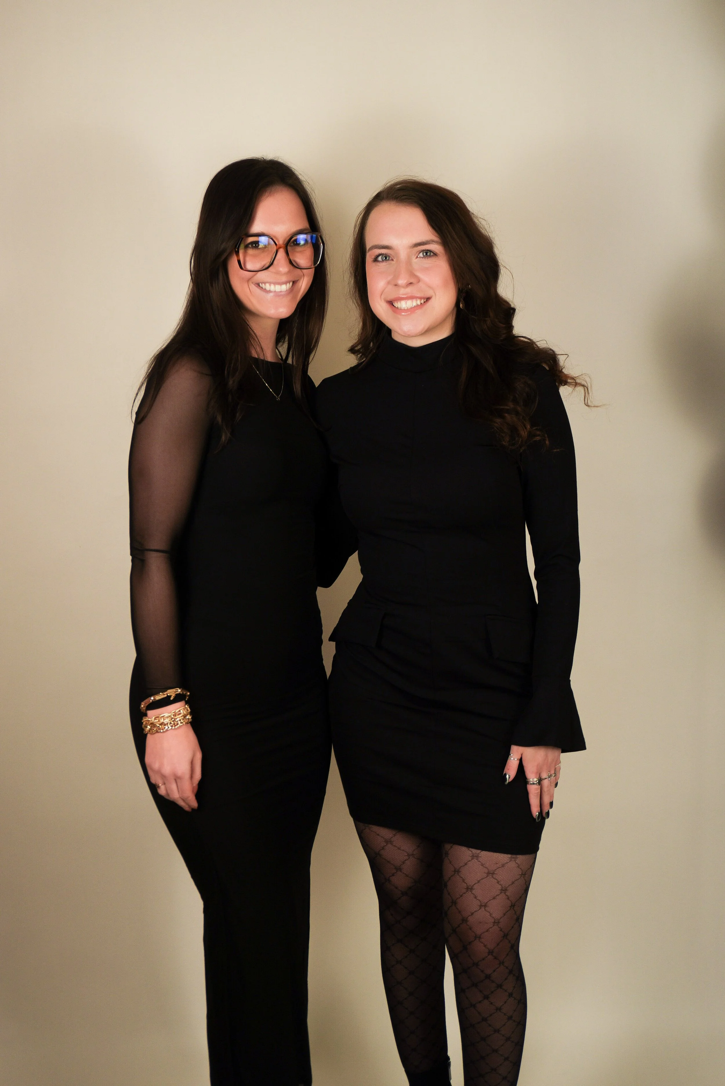 Two women standing close together, smiling, wearing black outfits, against a light-colored background.