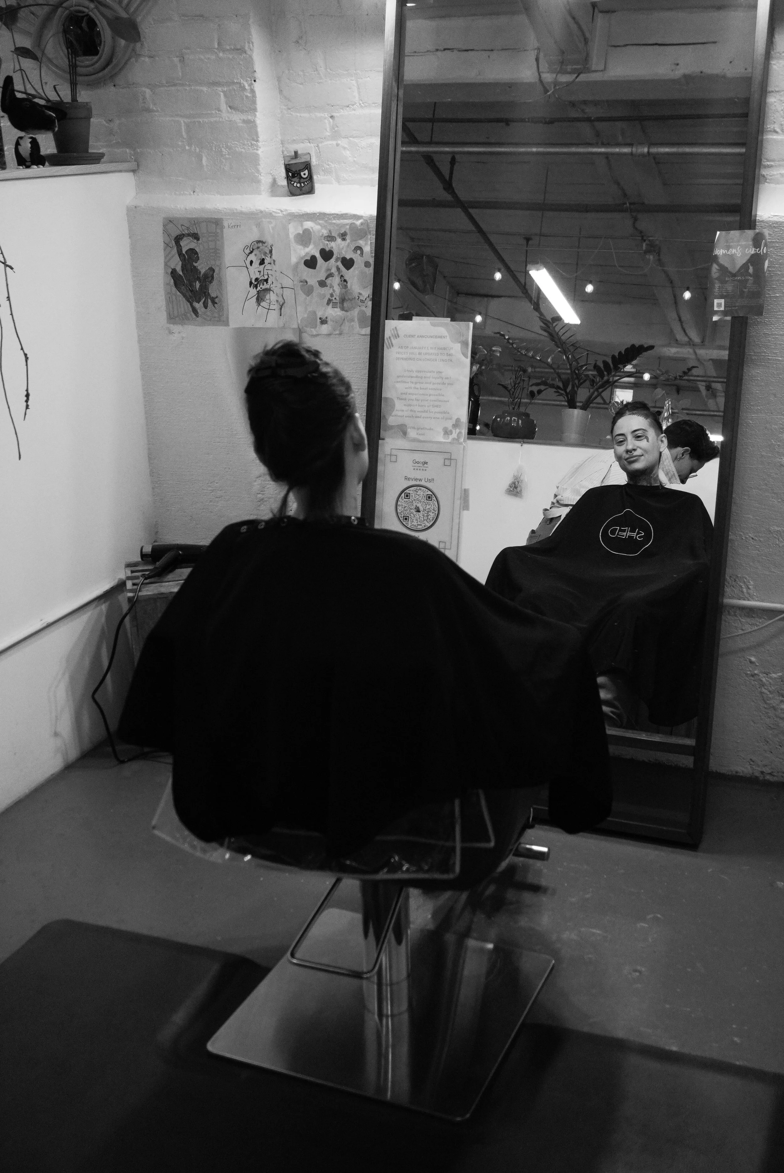 A woman getting her hair cut at a salon, sitting in a chair facing a mirror with her back to the camera, and a hairdresser working behind her.