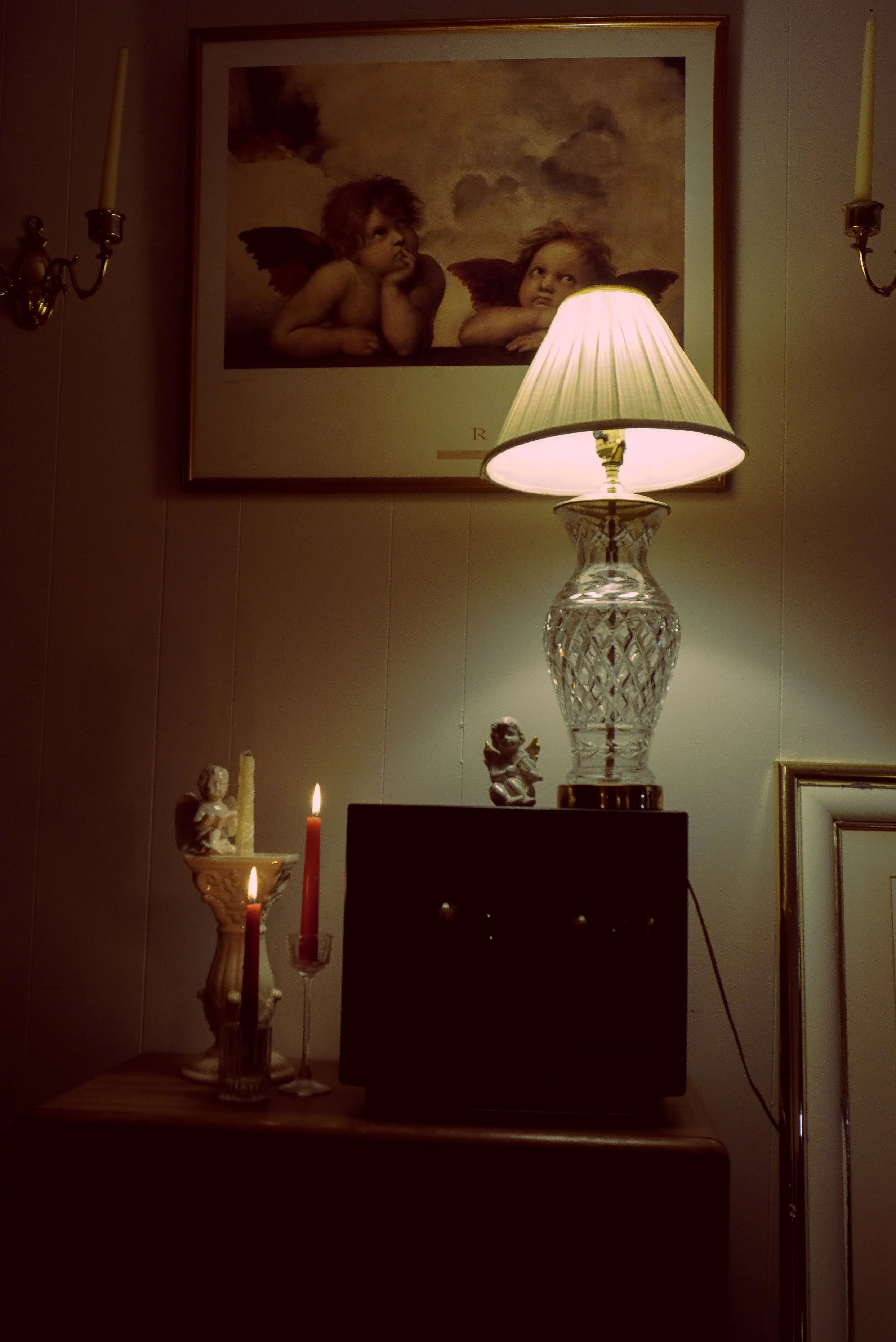 A lit table lamp with a glass base and a pleated shade, placed on a wooden surface. Behind the lamp, there is a small angel figurine and candles in candle holders. On the wall above, a framed painting depicts two cherub-like children with wings, resting their chins on their hands.