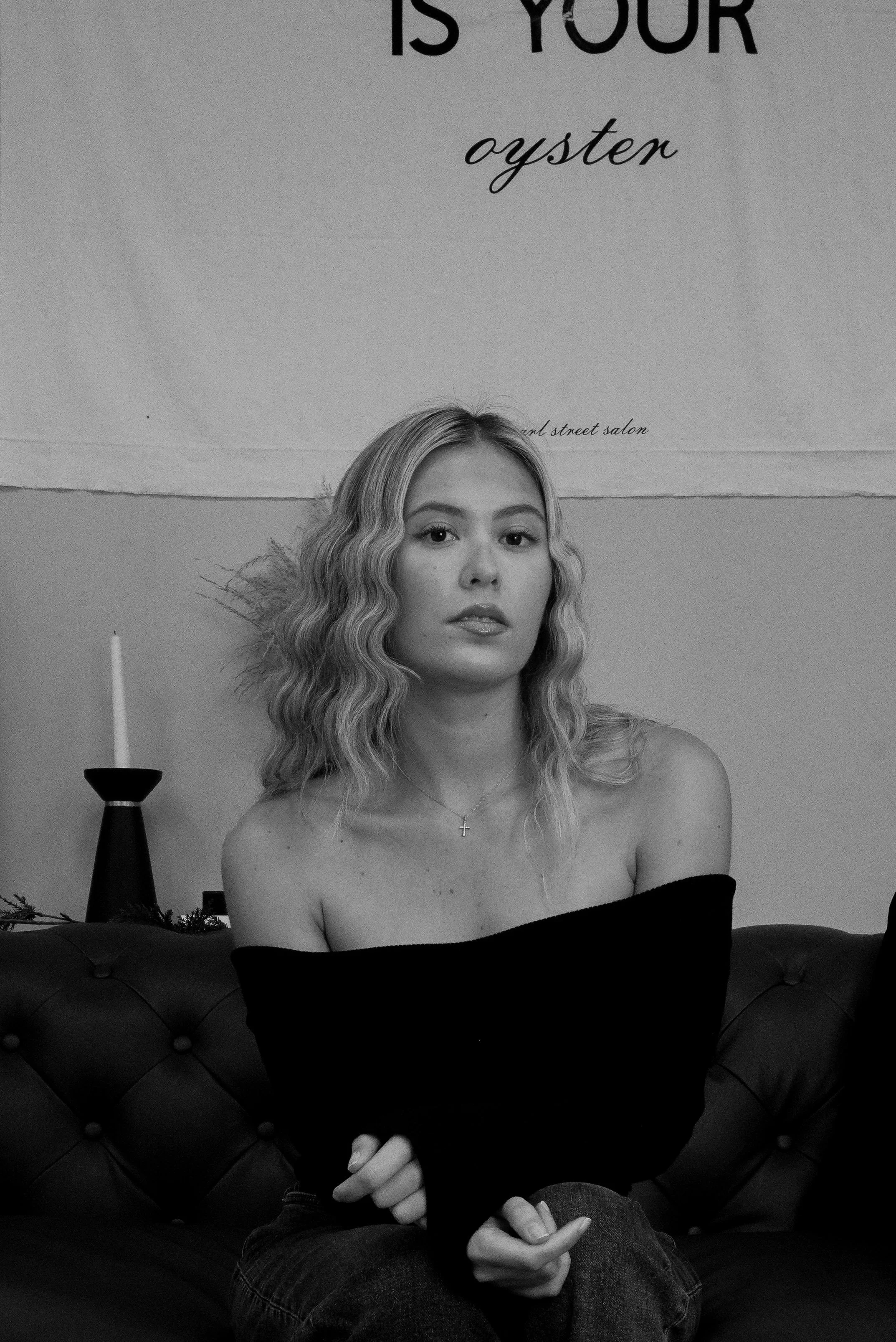 A young woman with wavy blonde hair sits on a tufted dark sofa, wearing an off-the-shoulder black top and a cross necklace, in front of a wall with a sign that reads 'IS YOUR oyster' and a candle in a holder on a side table.