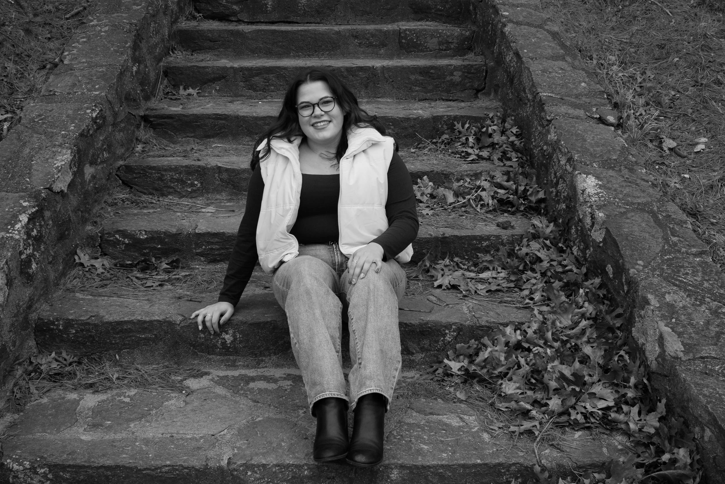 A woman with glasses sitting on outdoor stone steps, smiling, wearing a vest, jeans, and boots, with fallen leaves on the ground beside her.