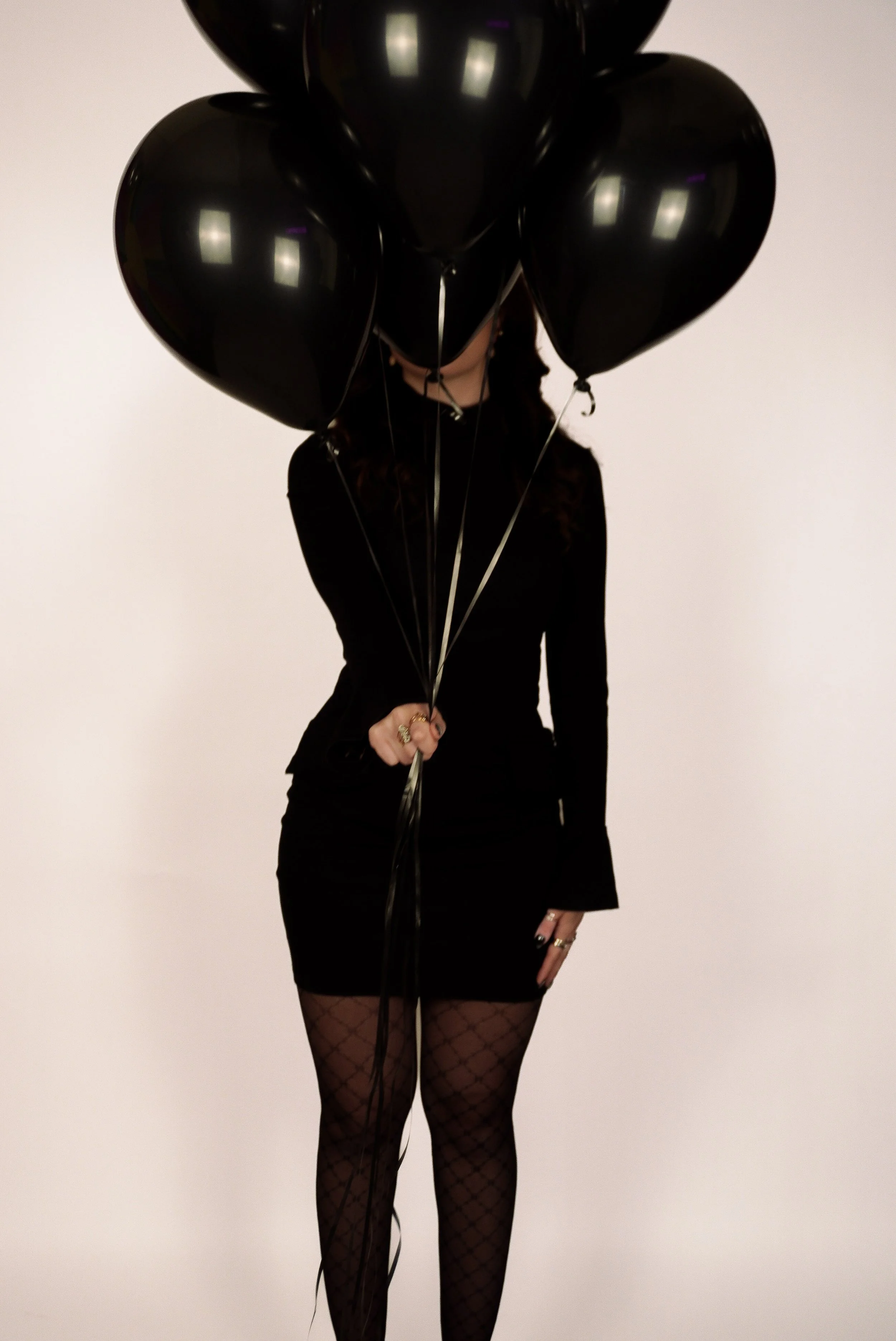 Person in black outfit holding several black balloons in front of a plain white background.