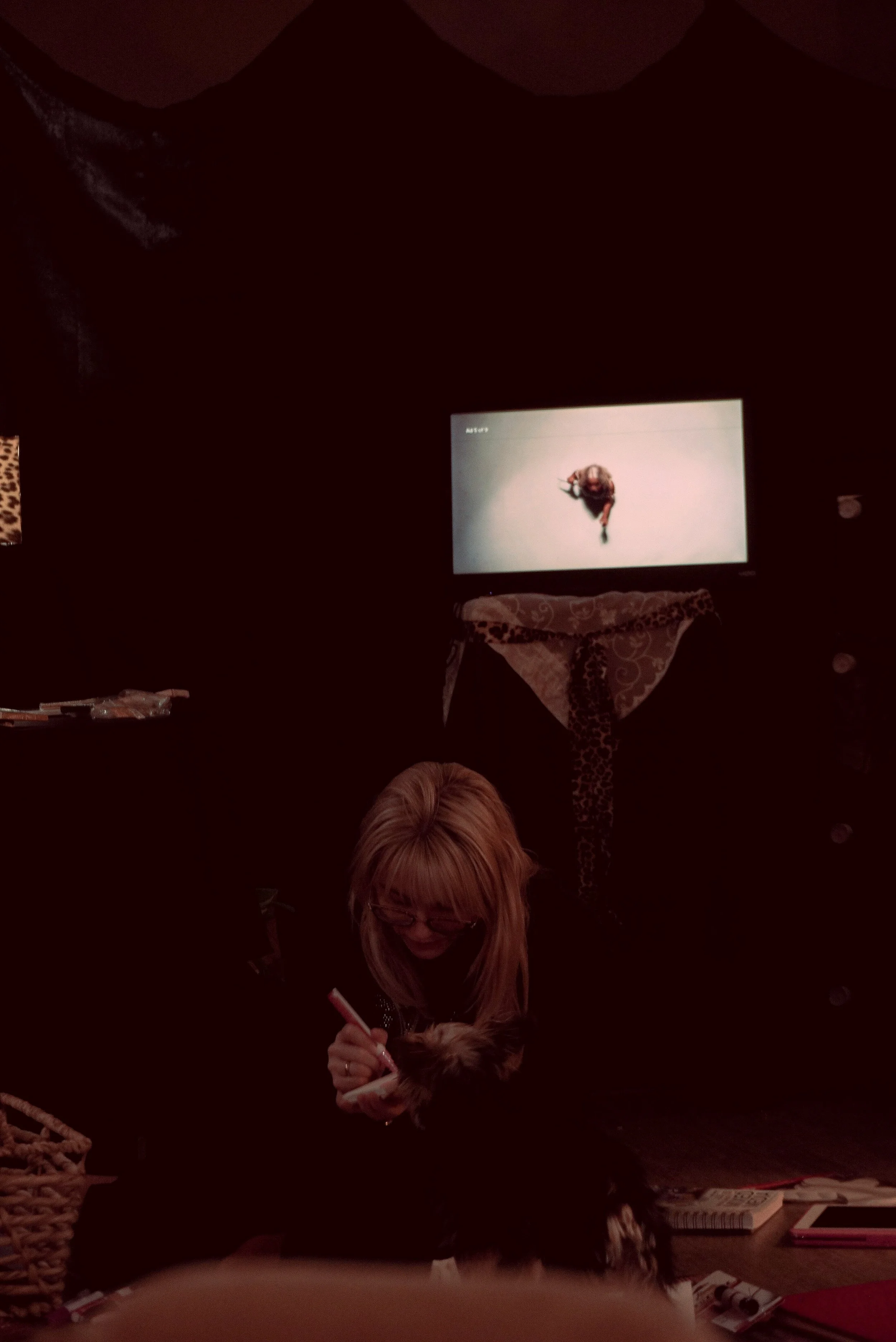 A woman with blonde hair and glasses sitting on the floor, writing or drawing on a small board or notepad, with a small fluffy dog on her lap, in a dimly lit room with a television on the wall showing an animated character.