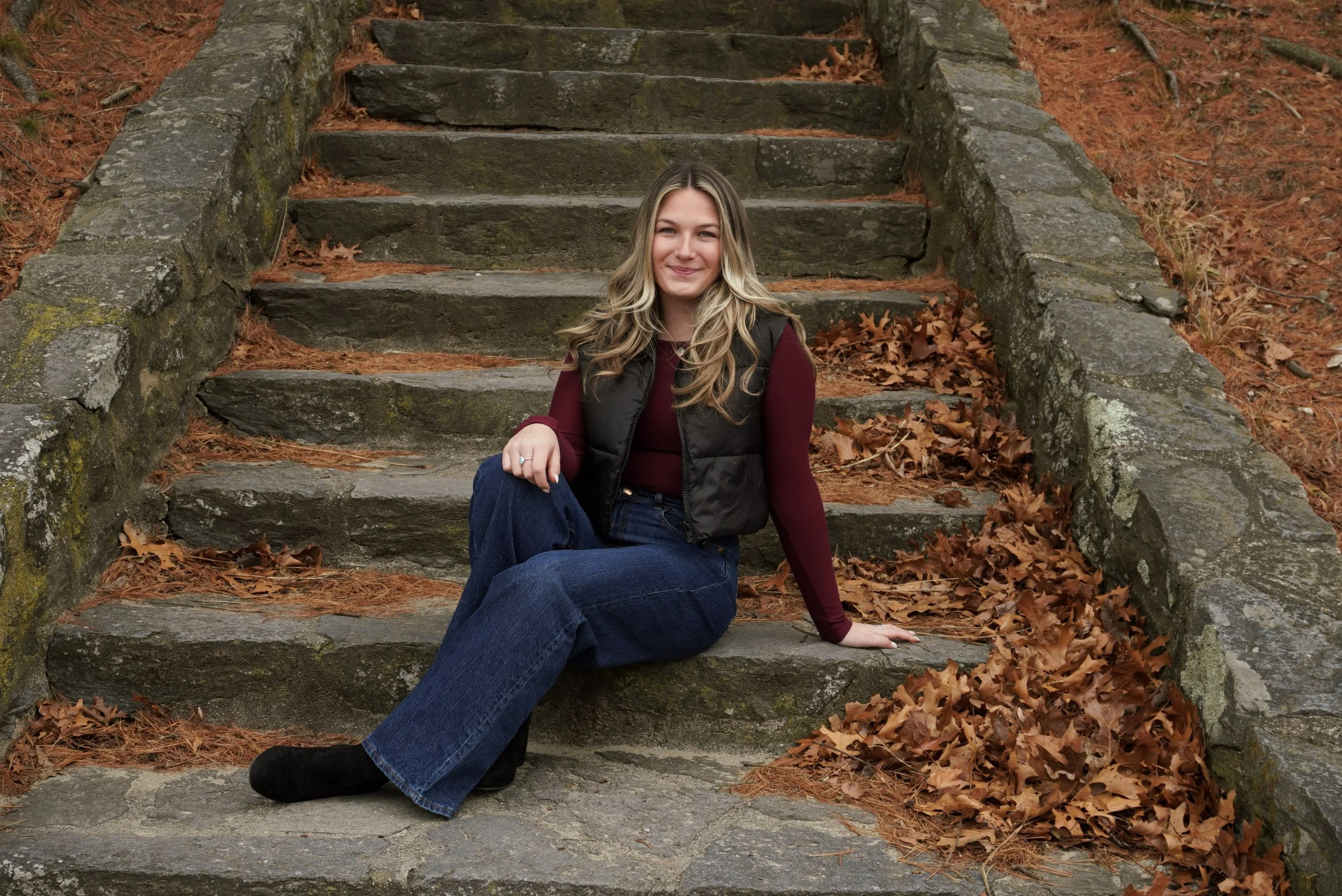 A young woman with long blonde hair sitting on stone steps outdoors during autumn, surrounded by fallen brown leaves, wearing a maroon long sleeve shirt, black vest, blue jeans, and black boots, smiling at the camera.