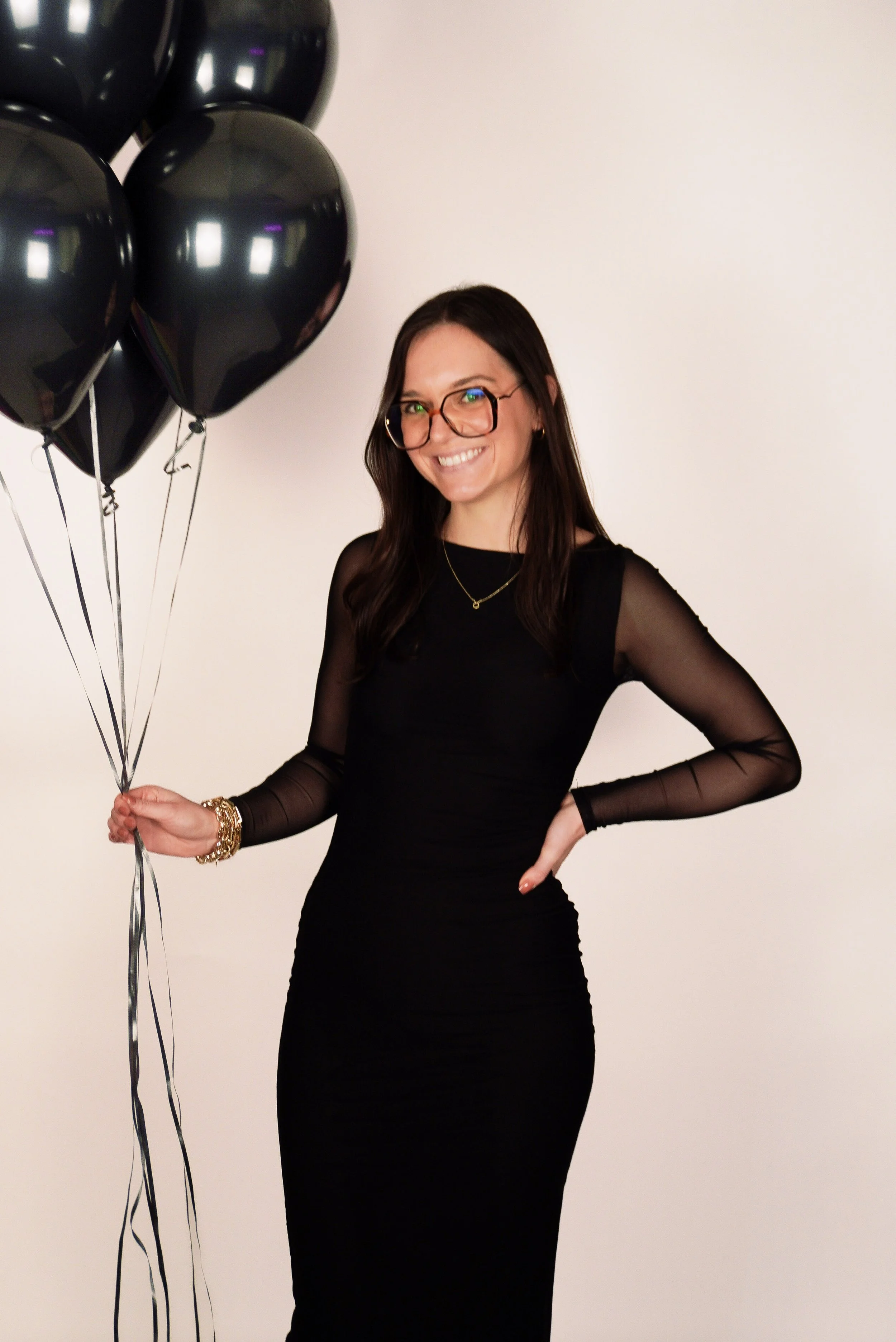 A woman smiling, wearing glasses and a black dress, holding a bunch of black balloons, standing against a white background.