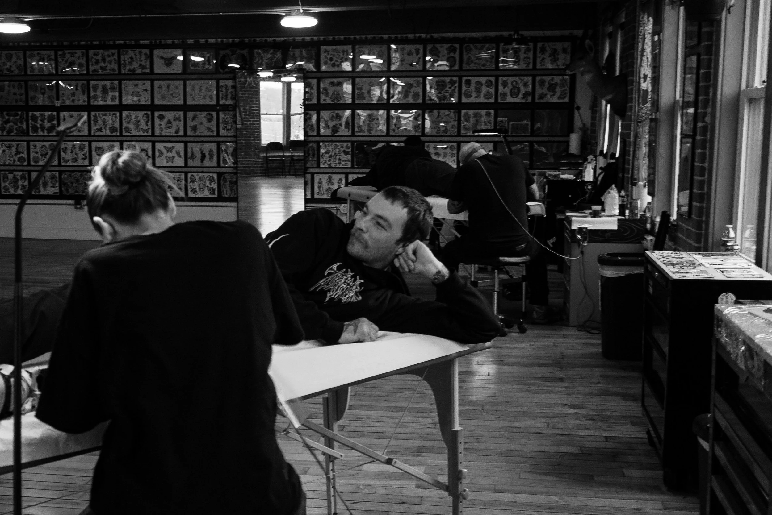 A man lying on a tattoo artist's table in a tattoo shop, looking at a woman seated across from him. The shop has tattoo flash artwork on the walls and a wooden floor.