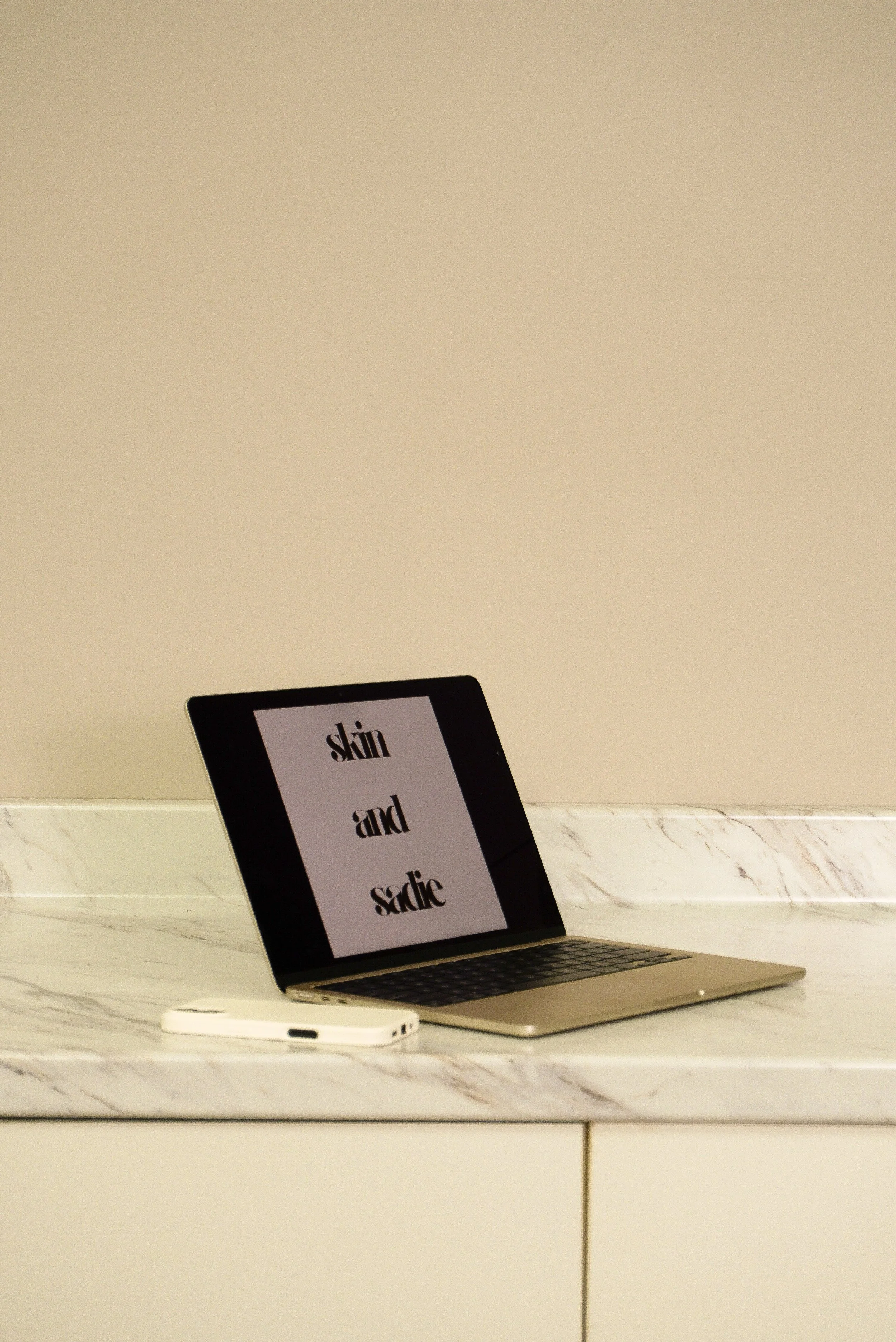 A laptop on a marble countertop displaying the words 'skin and saxie,' with a smartphone placed nearby.