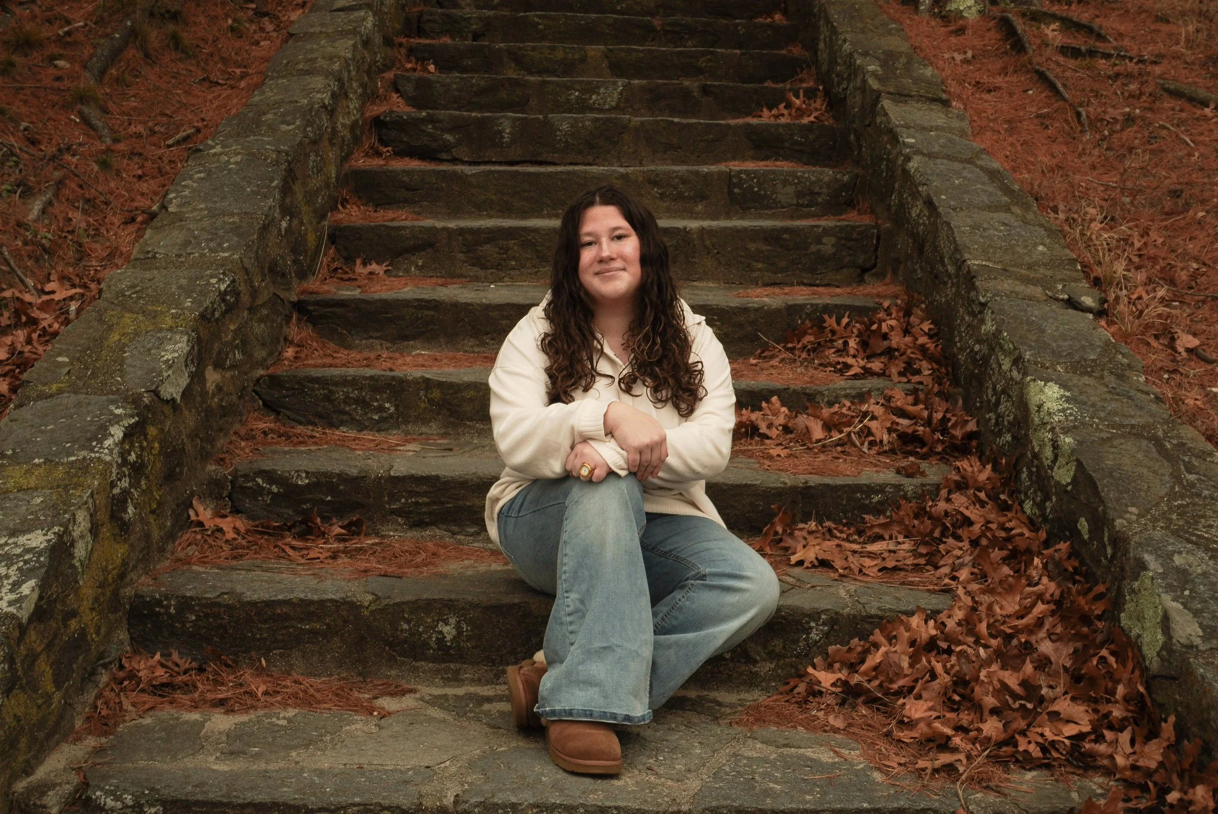 A woman with long, curly brown hair sitting on stone stairs covered with brown autumn leaves in a wooded area.