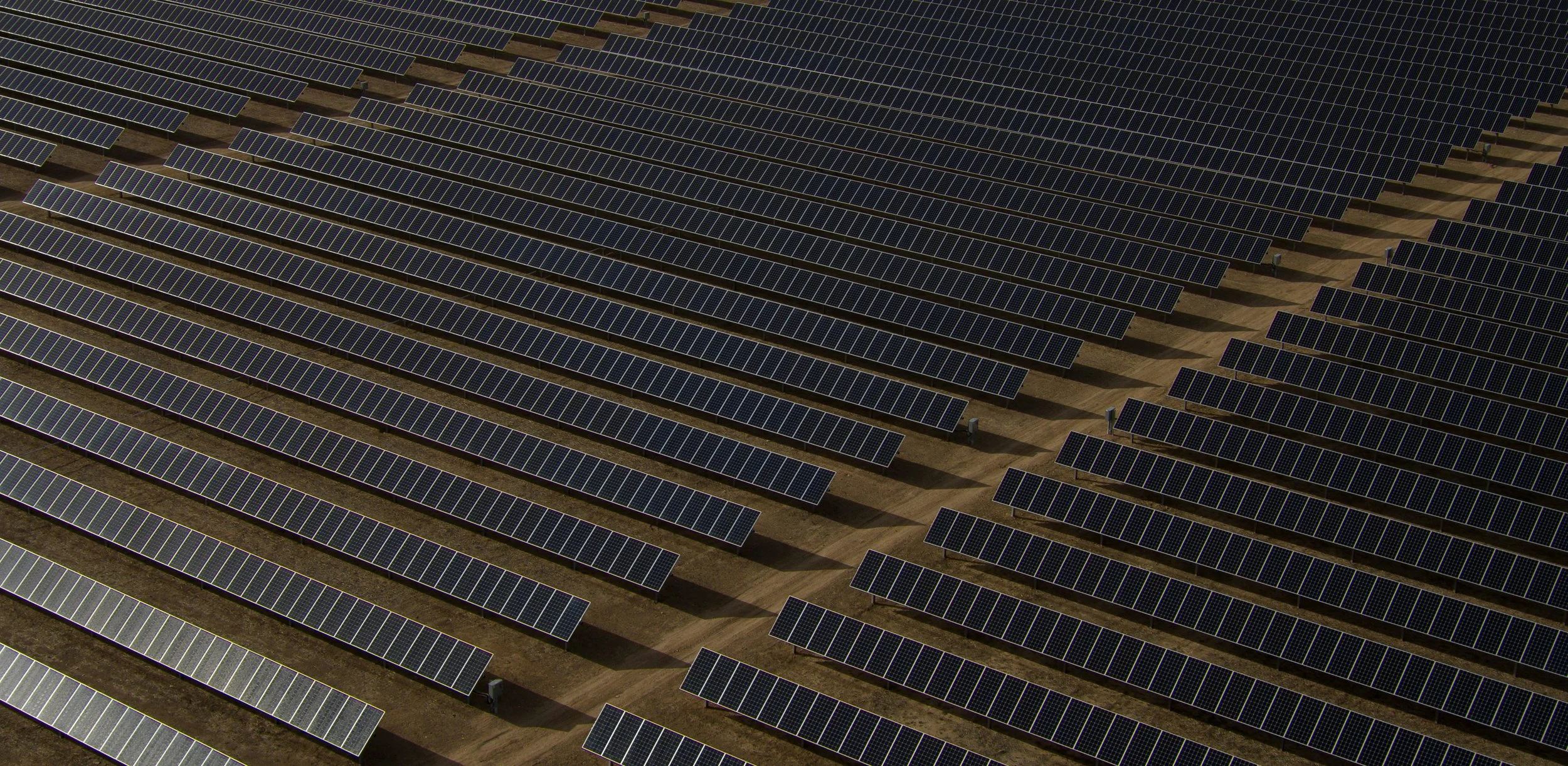 An aerial view of a large solar farm with numerous solar panels arranged in neat, parallel rows on the ground.