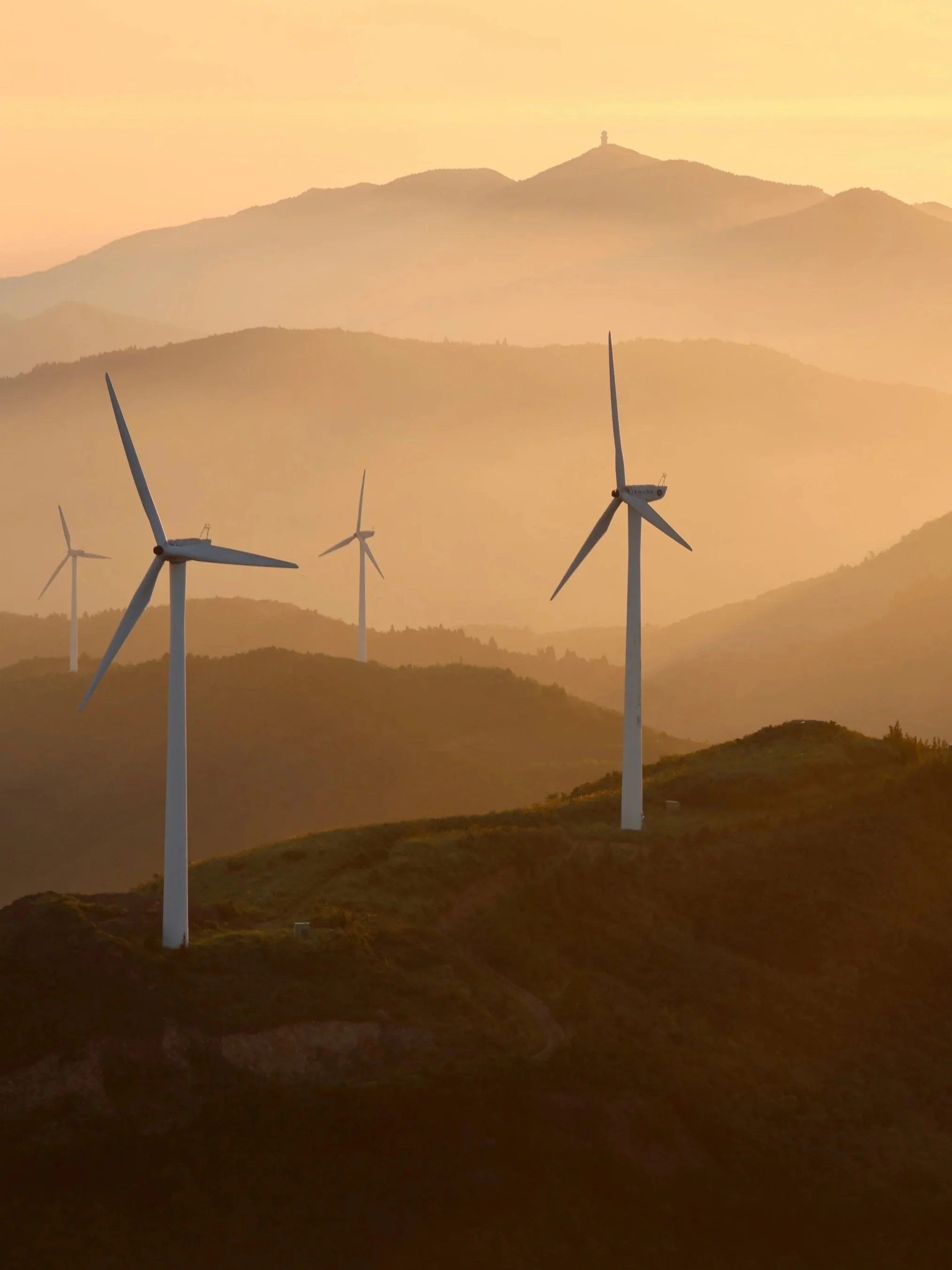Several wind turbines are on a hill during sunset, with mountains in the background.