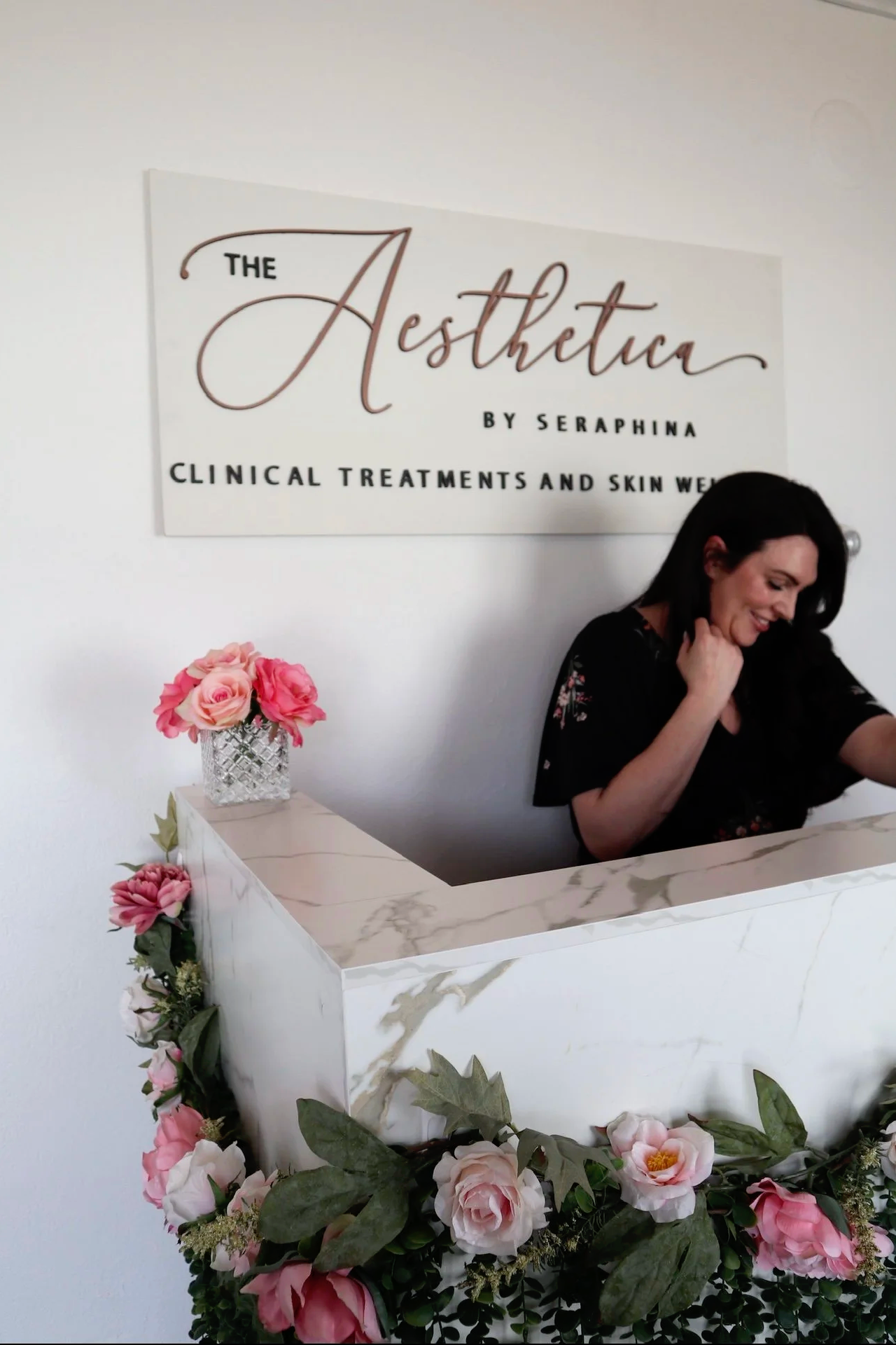Reception desk decorated with pink and white florals, and green foliage. The Aesthetica founder is standing behind it smiling.