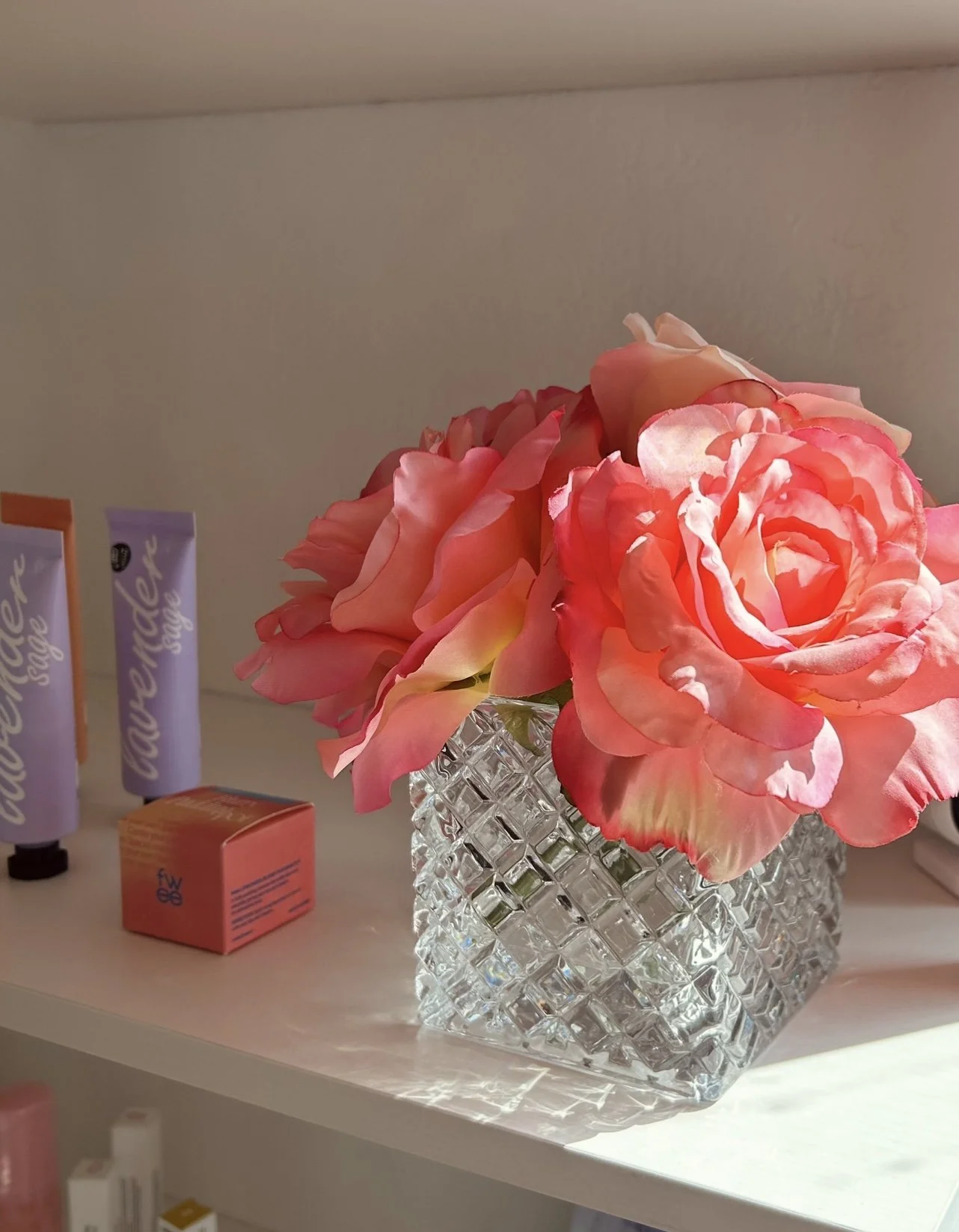 Pink and coral coloured roses in a textured crystal glass vase, on a white shelf.