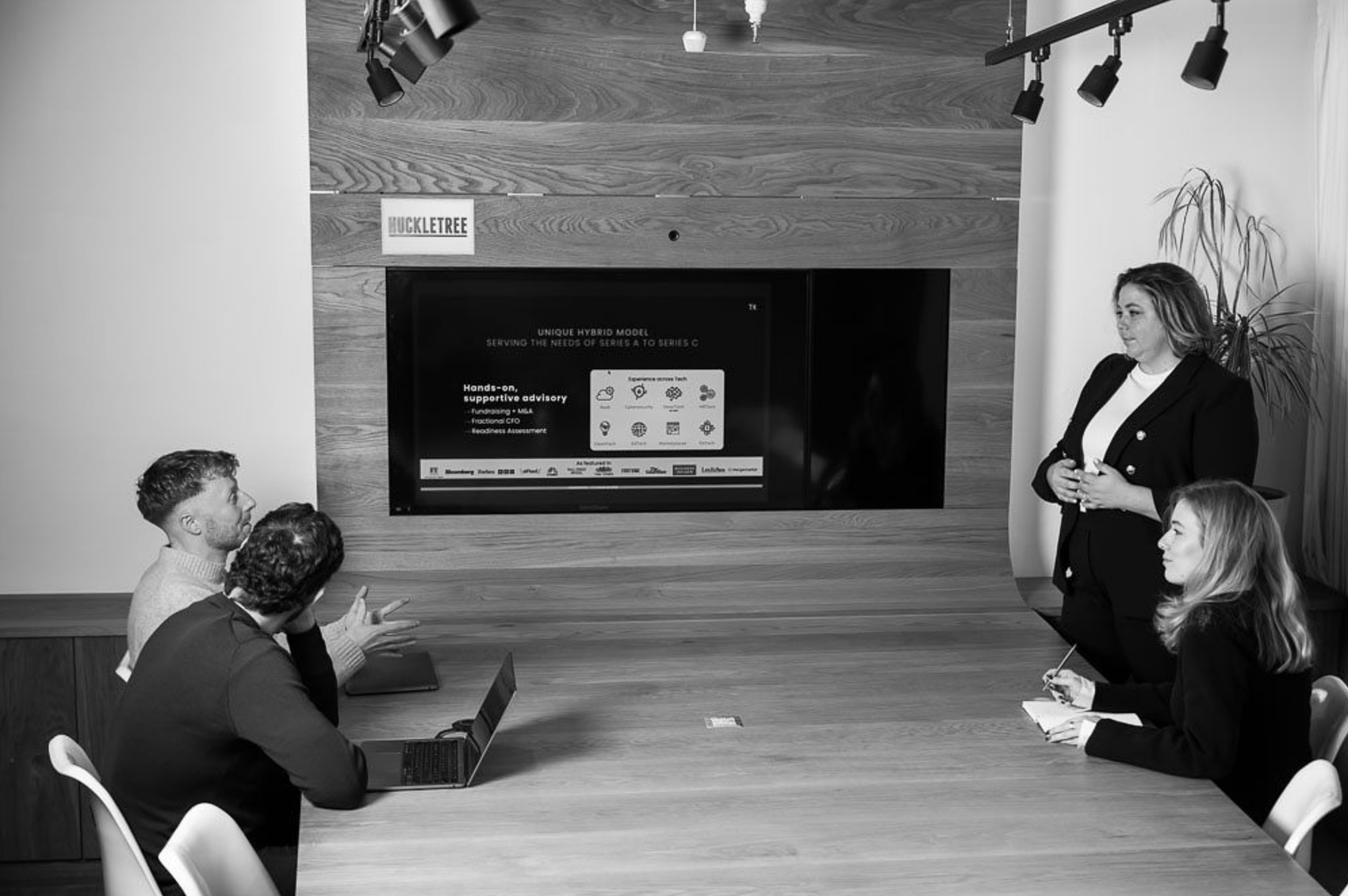 Group of four people in a business meeting or presentation in a conference room with a large monitor displaying a slide, and a woman speaking while three others are listening and taking notes.