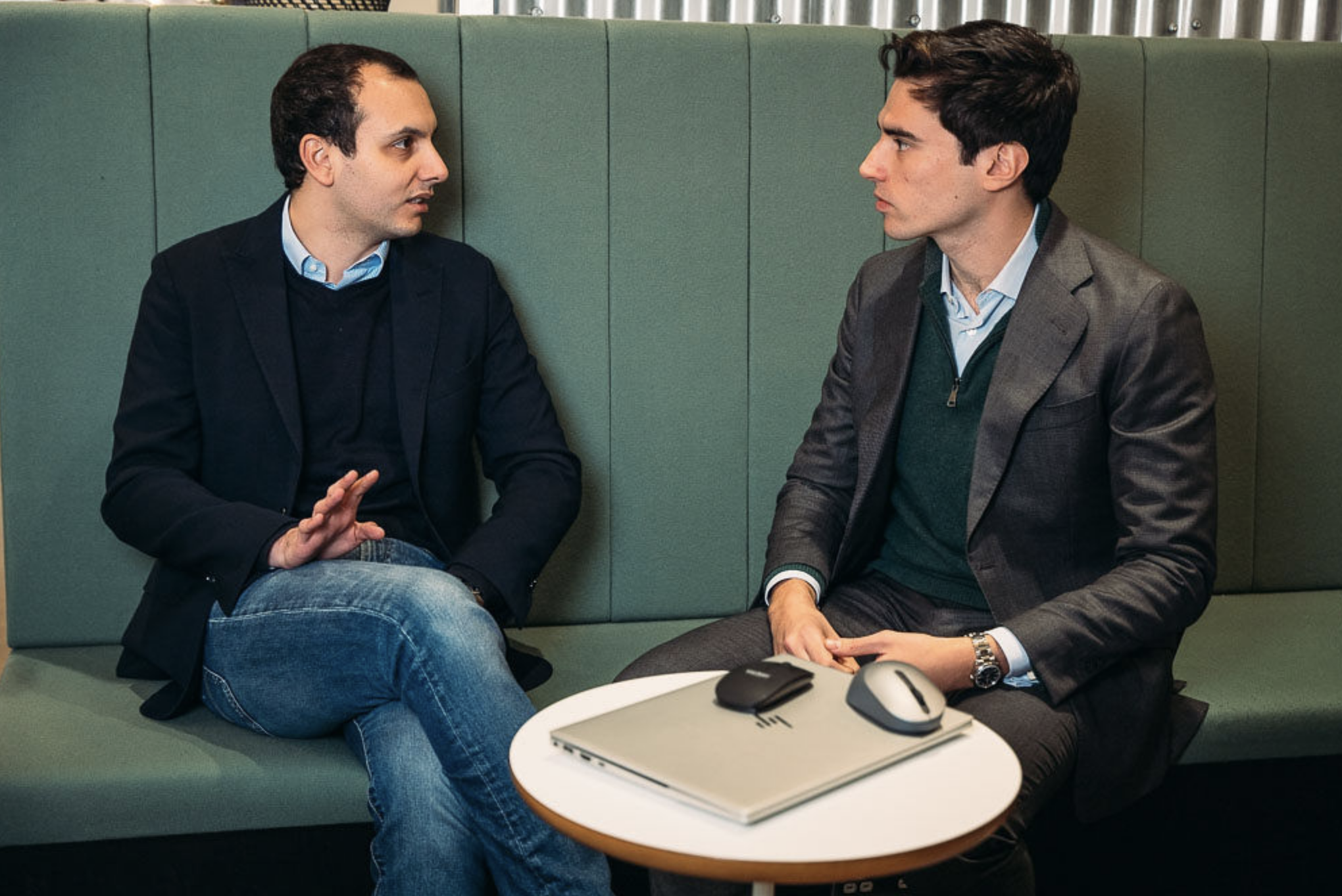 Two men sitting on a green booth seat having a serious conversation. One man is wearing a dark blazer and jeans, gesturing with his hand. The other man is in a grey suit, sitting with hands clasped, with a laptop, mouse, and another device on the small white round table in front of them.