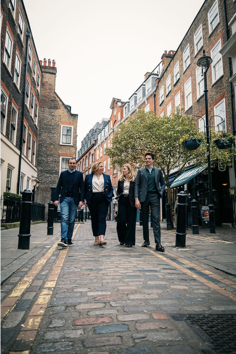 Four business professionals walking down a cobblestone street in an urban area, surrounded by multi-story brick buildings and trees.