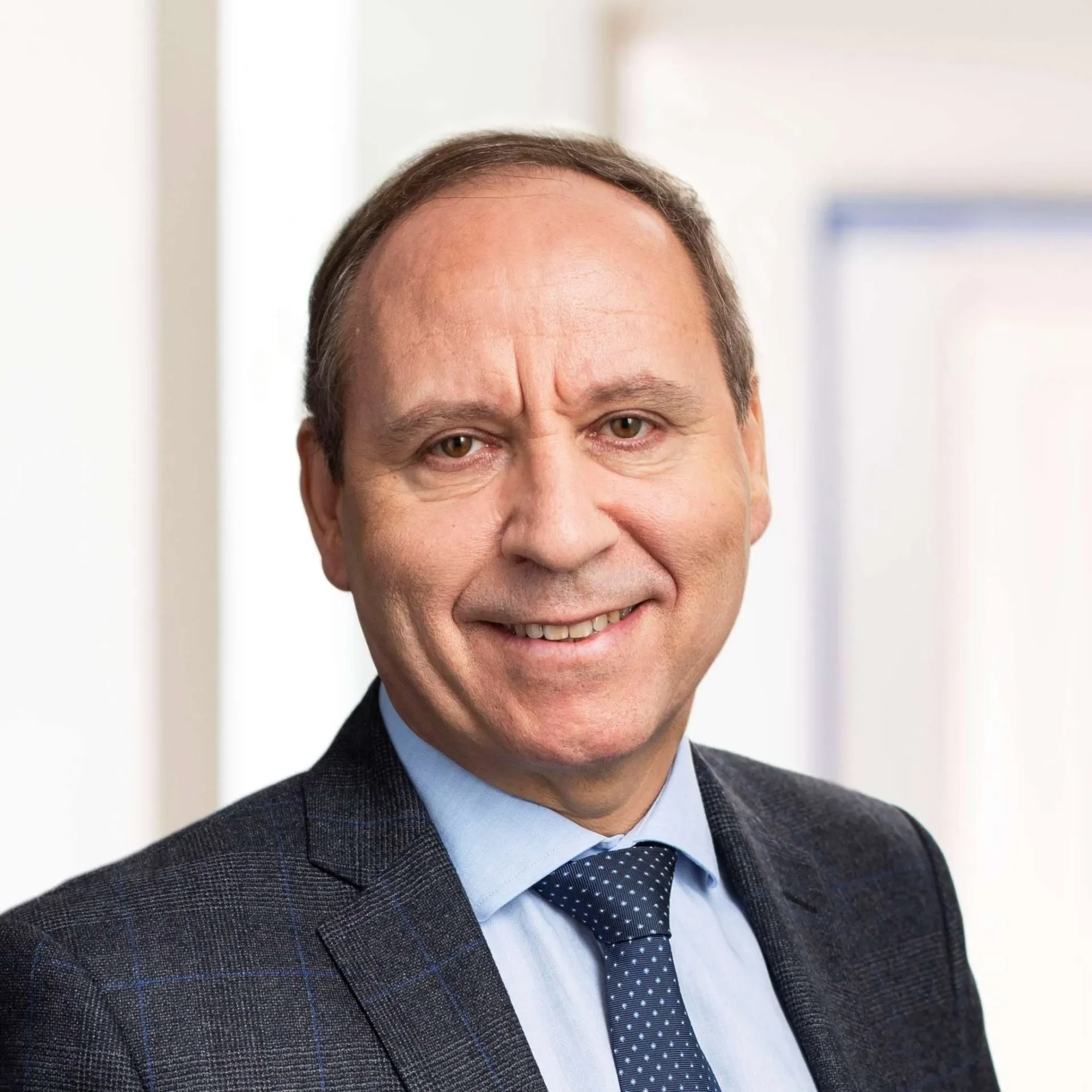 A professional headshot of a middle-aged man in a suit, smiling, indoors with blurred background.