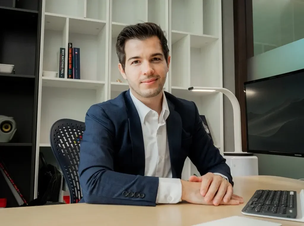 A young man in a business suit sitting at a desk in an office, smiling with a bookshelf and computer monitor behind him.