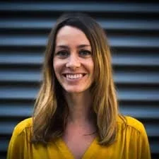 A woman with shoulder-length brown hair, wearing a yellow top, smiling, with a dark, slatted background.