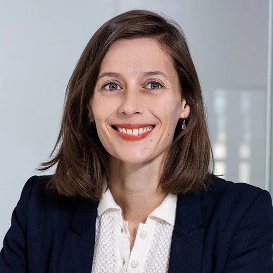 A woman with shoulder-length brown hair wearing a white blouse and a dark blazer, smiling in a professional setting.