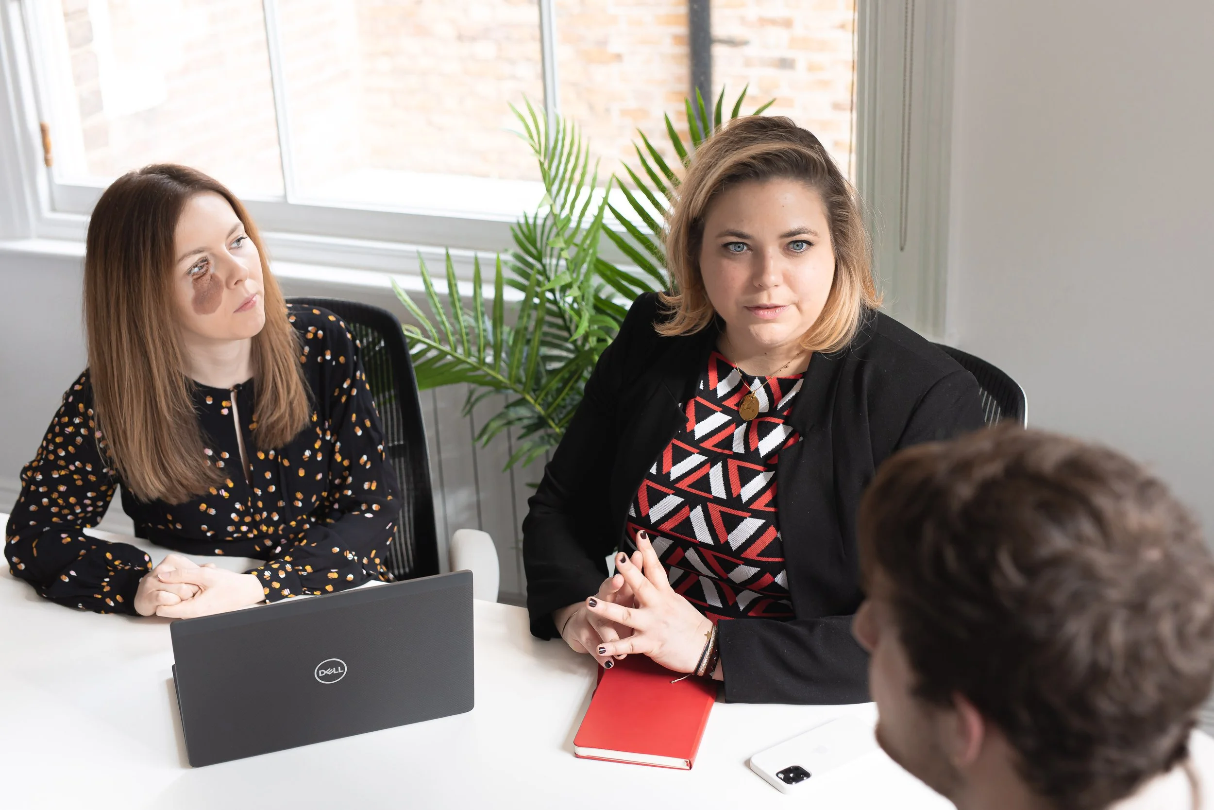Three people engaged in a discussion at a meeting table in a modern office with large windows and a potted plant. One woman is facing the camera, wearing a black jacket over a patterned top, with a red notebook and a smartphone on the table. The other woman has brown hair with dark spots on her face and is looking to the side, wearing a black blouse with gold dots. The man, whose back is to the camera, is talking to the woman facing dawn.
